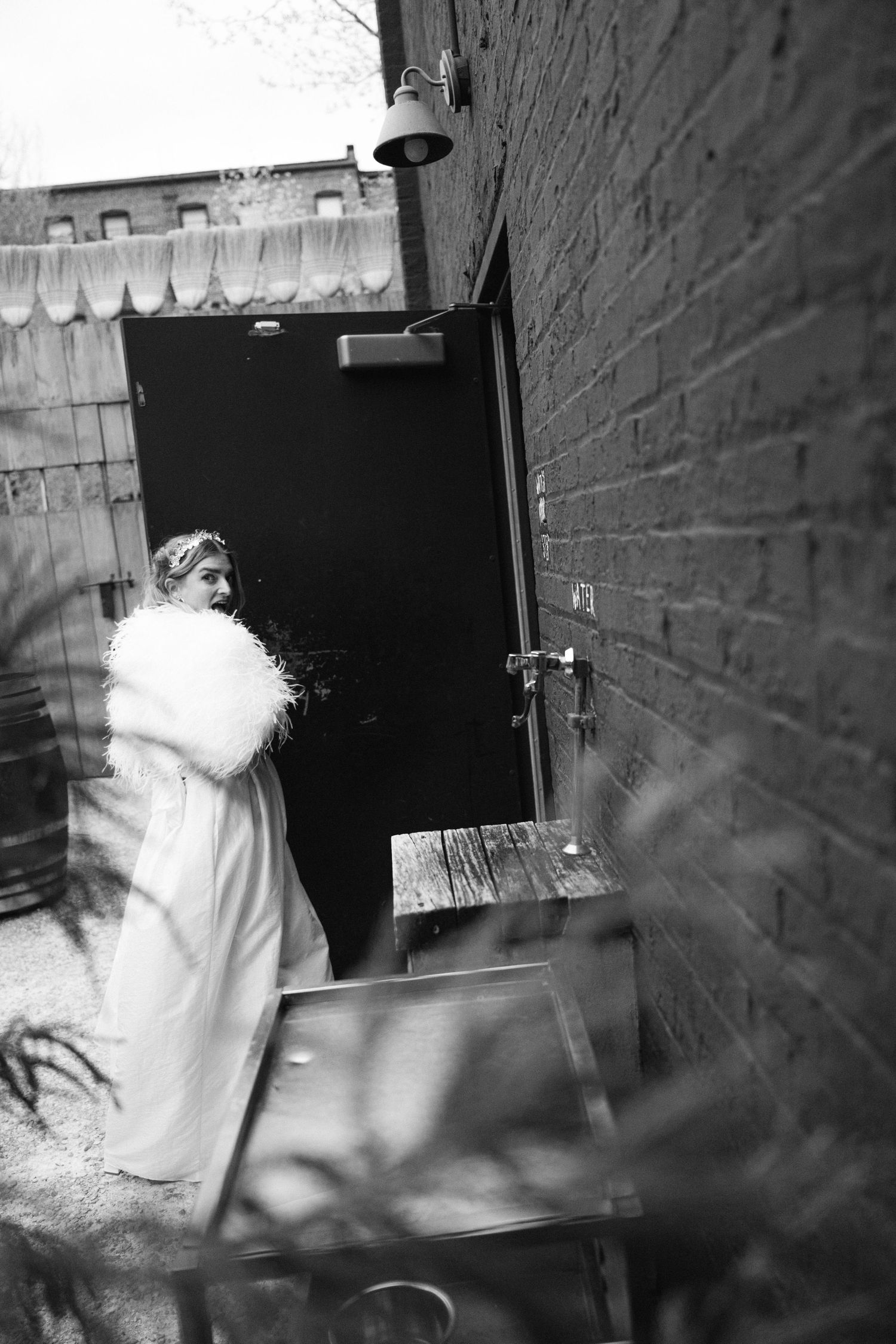 A person in a white dress and furry wrap stands in a doorway of a brick building.