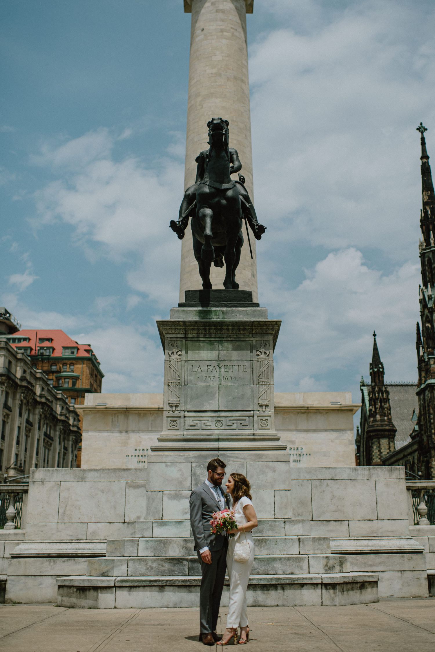 Couple poses beneath towering monument and classical architecture in romantic urban setting.