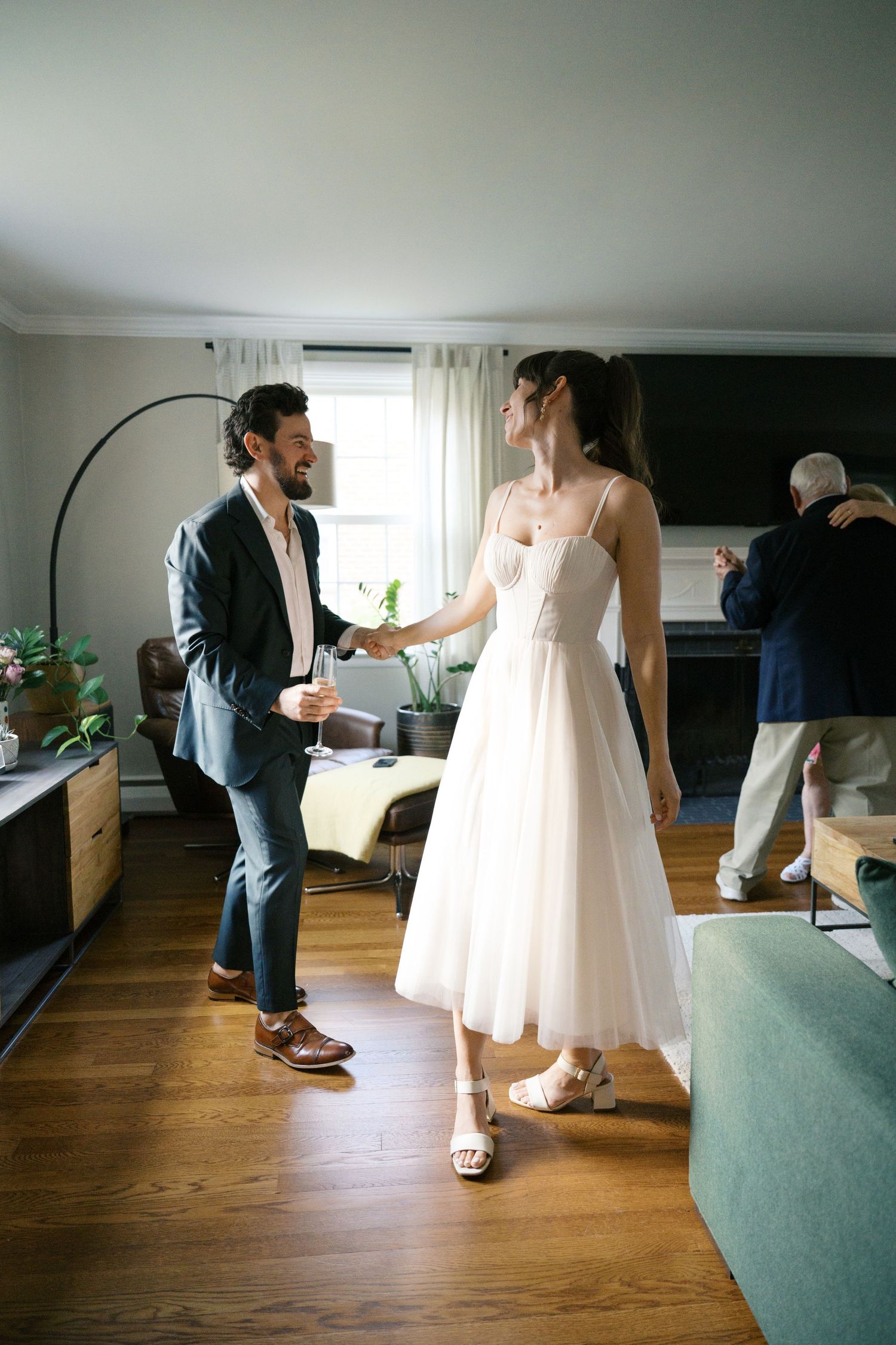 A couple shares a sweet moment in a living room, her in a white dress and him in a navy suit.
