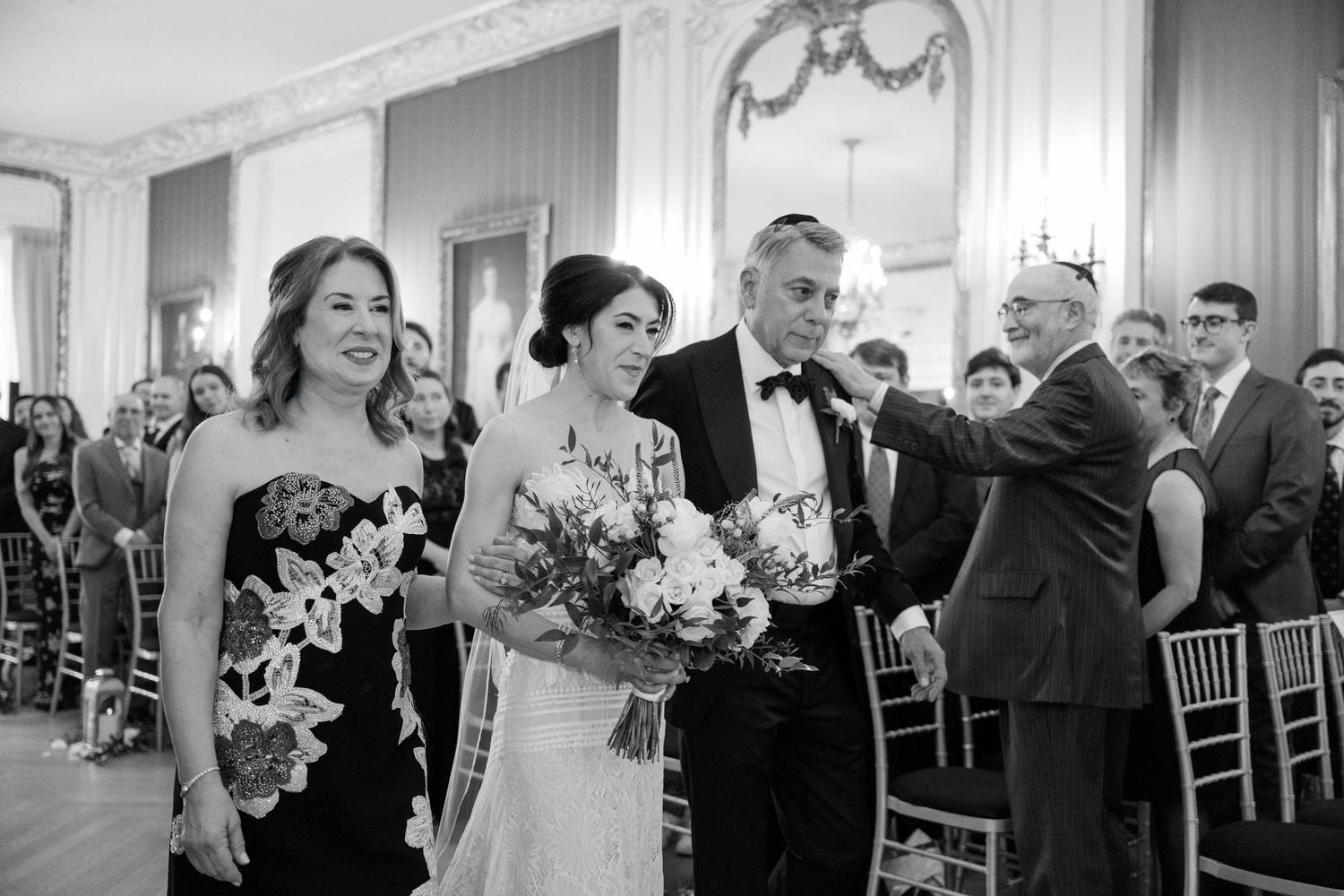 Wedding ceremony processional in a grand ballroom with ornate white decor.