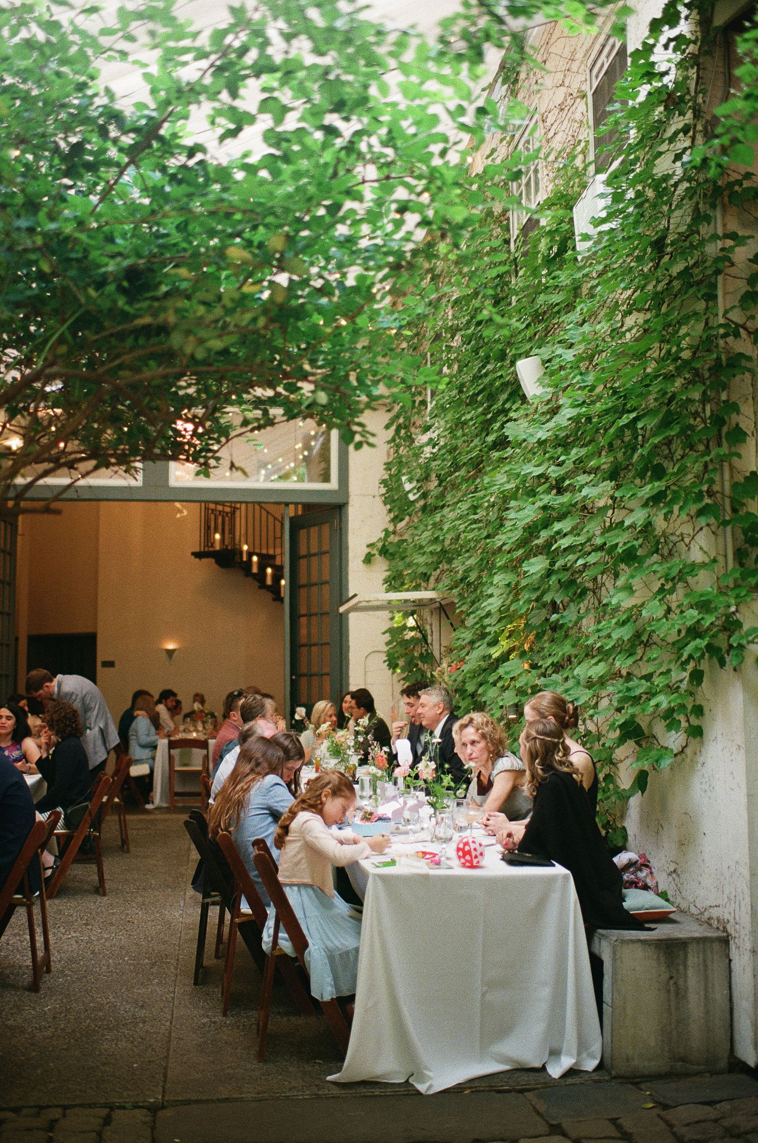 Elegant outdoor wedding reception with white-clothed tables under lush green foliage and string lights.