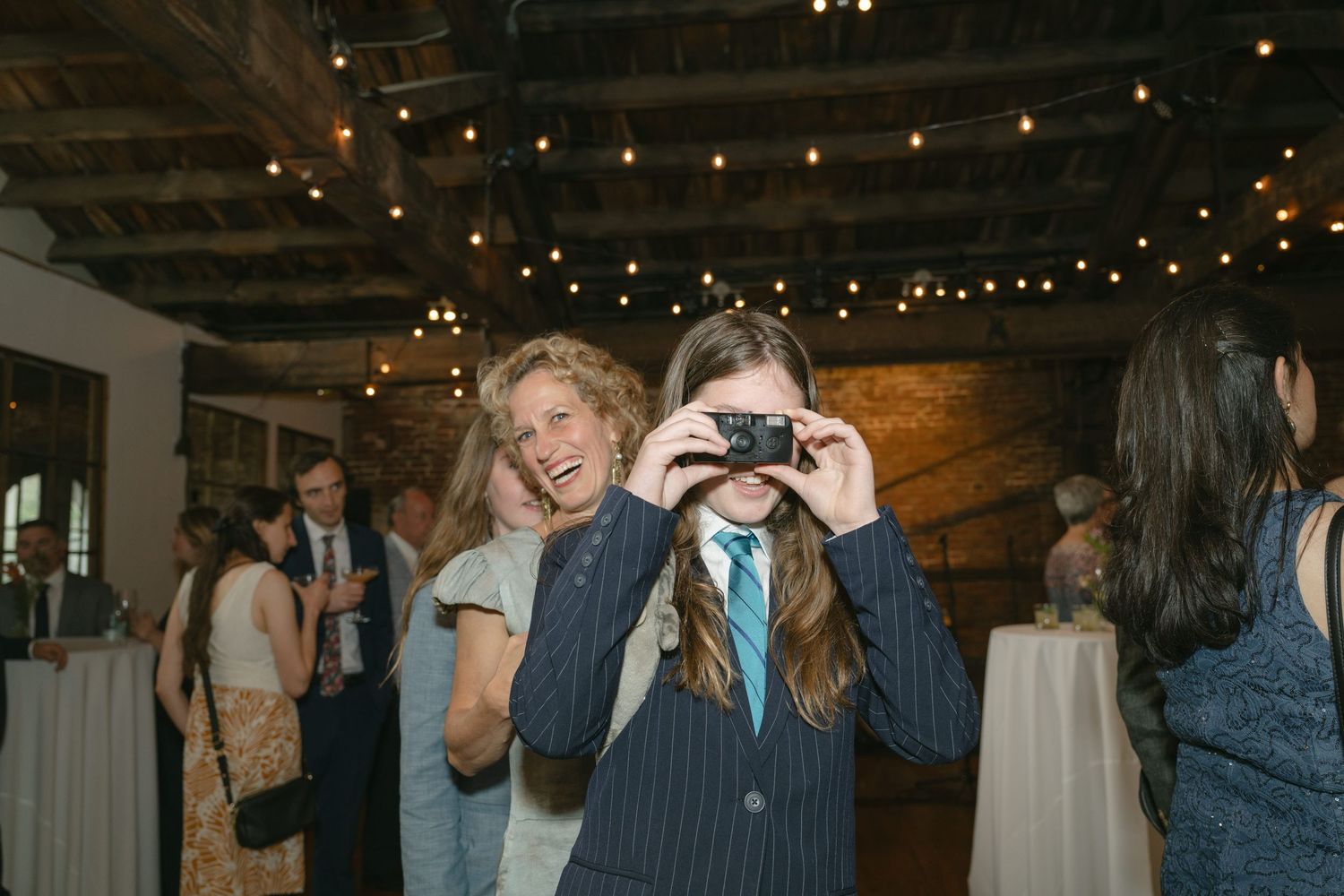 Indoor event with string lights overhead as someone takes a photo with a camera in a rustic venue space.