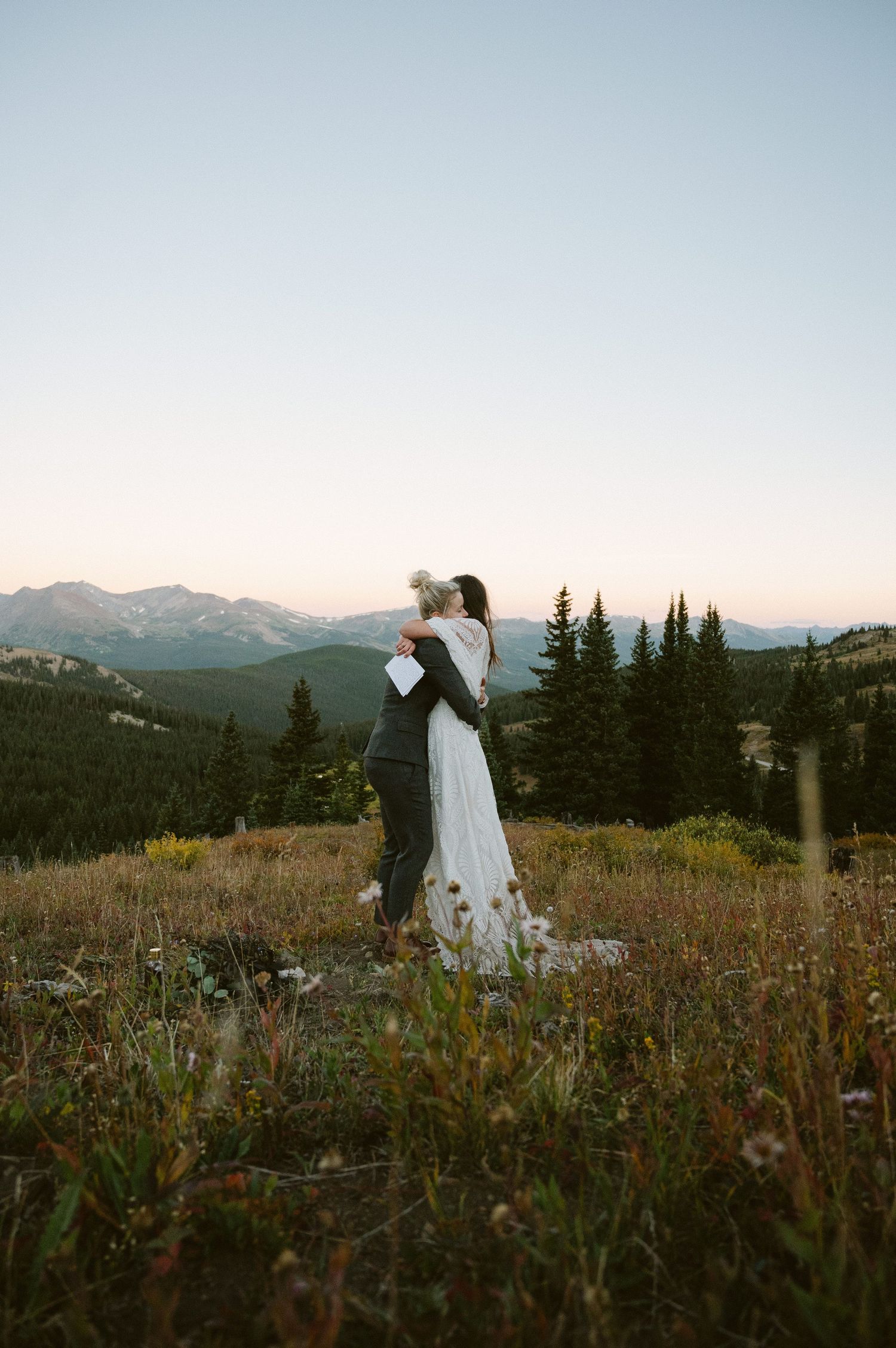 Bride and groom embrace in a mountain meadow at sunset, surrounded by evergreen trees and scenic peaks.