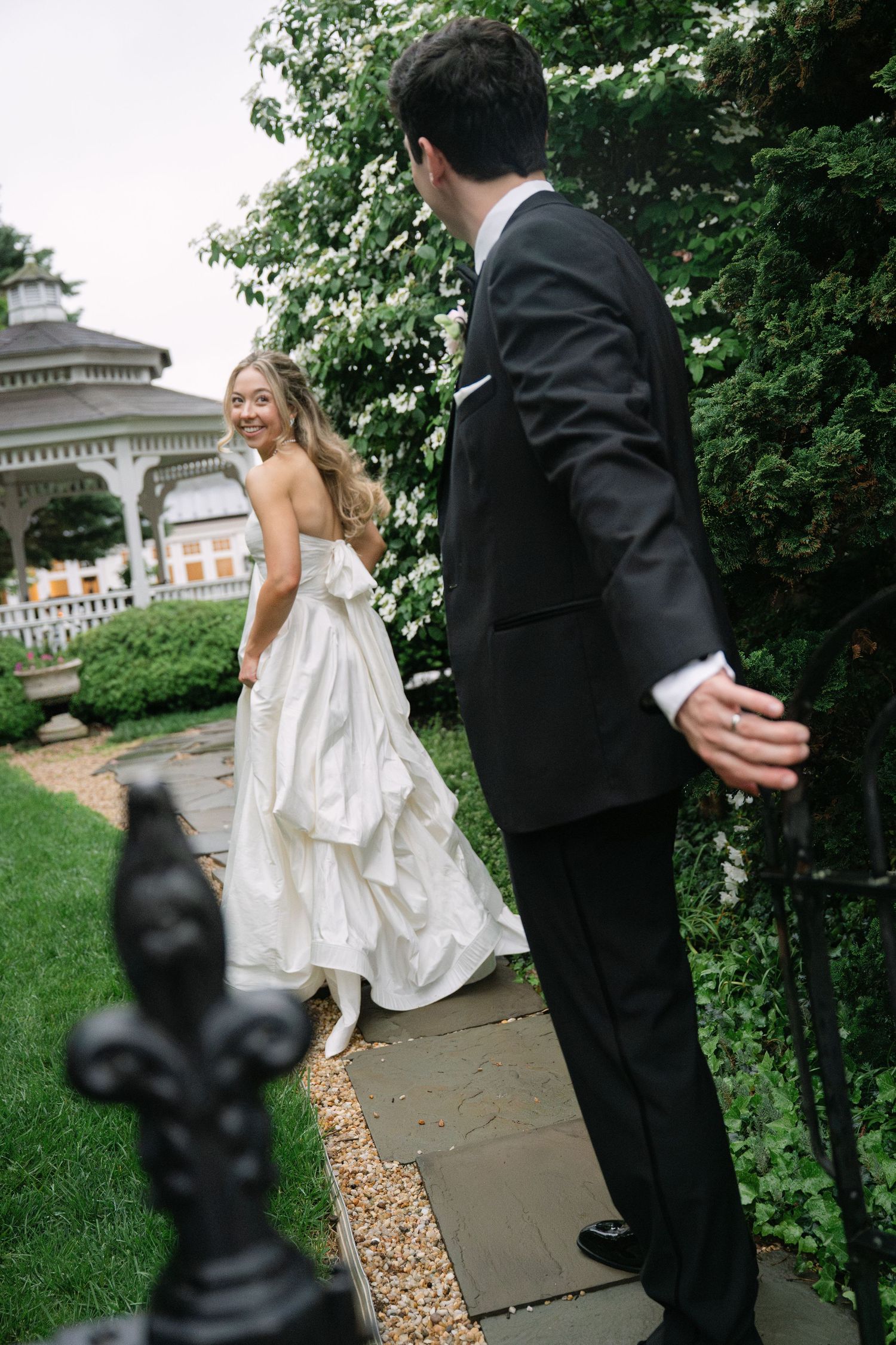 Bride in strapless white gown smiles at groom in black suit on garden path near gazebo.