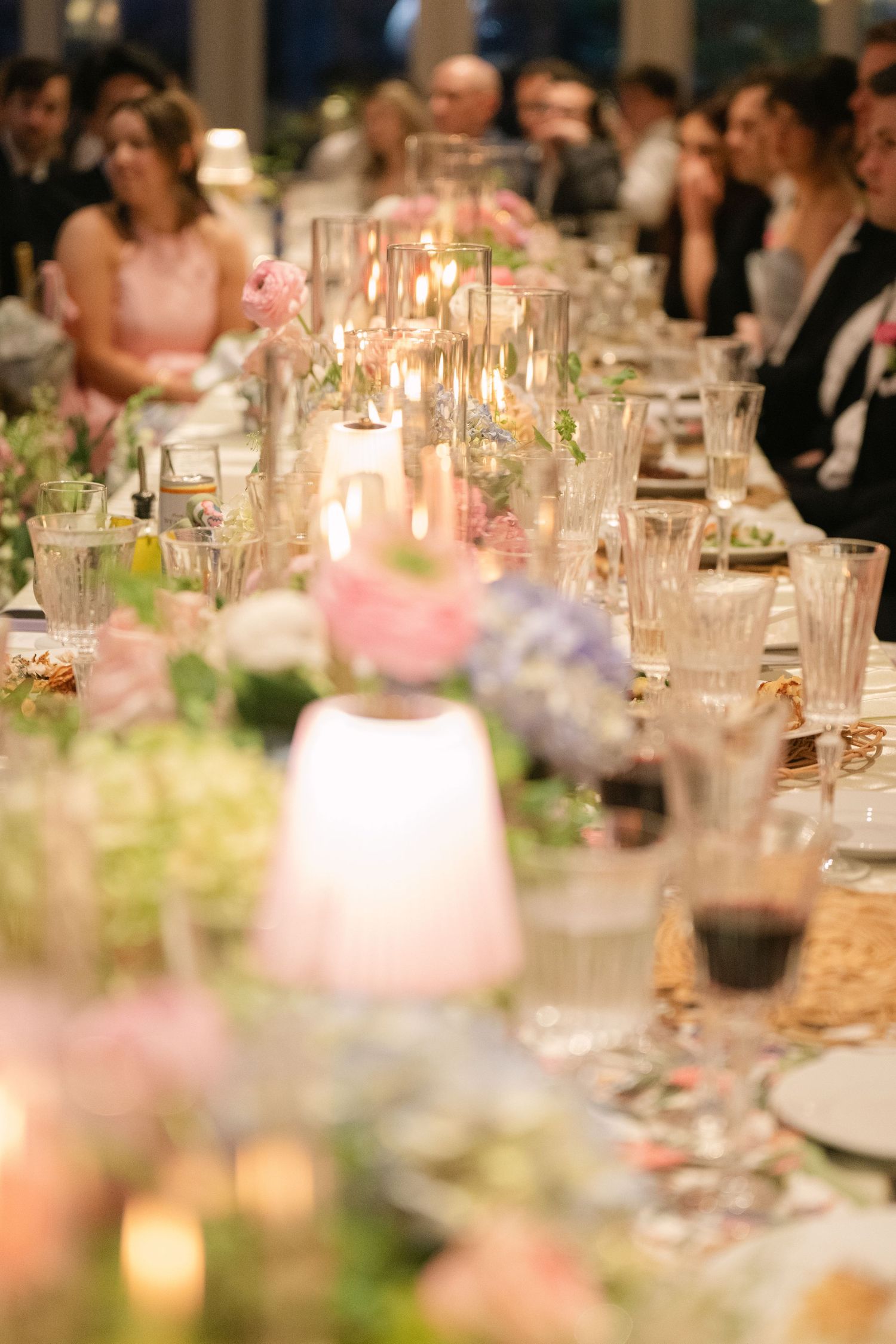 Elegant wedding reception table with crystal glasses, pink floral centerpieces, candles, and guests seated in background.