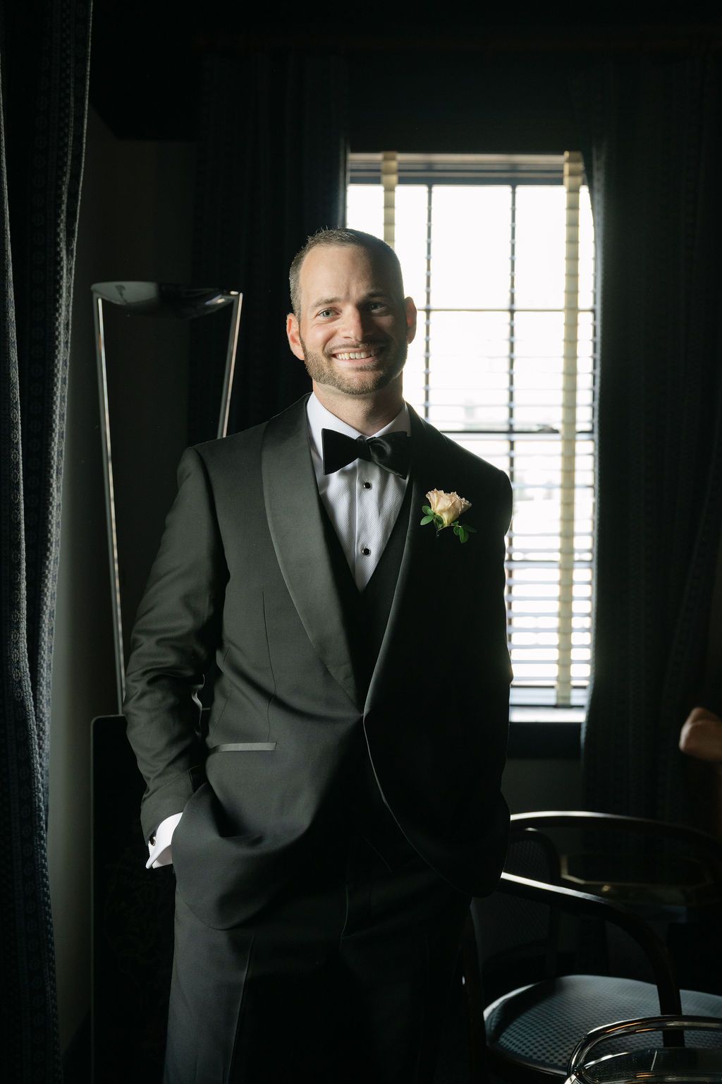 A groom in a classic black tuxedo stands by a window in a dimly lit room with natural light streaming in.