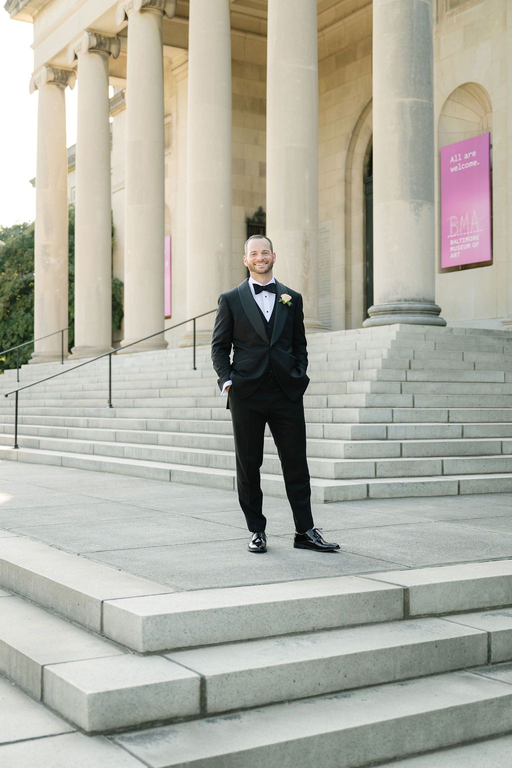 A person in a black tuxedo stands on grand stone steps with classical columns in the background.