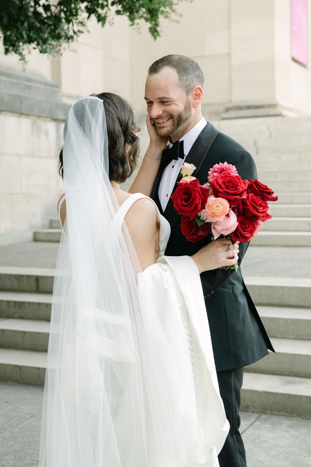 A romantic wedding moment captured on stone steps with a flowing white veil and vibrant red rose bouquet.