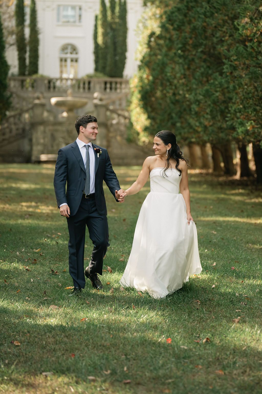 A couple playfully runs together on a grassy lawn near a historic stone building at Strong Mansion on Sugarloaf Mountain