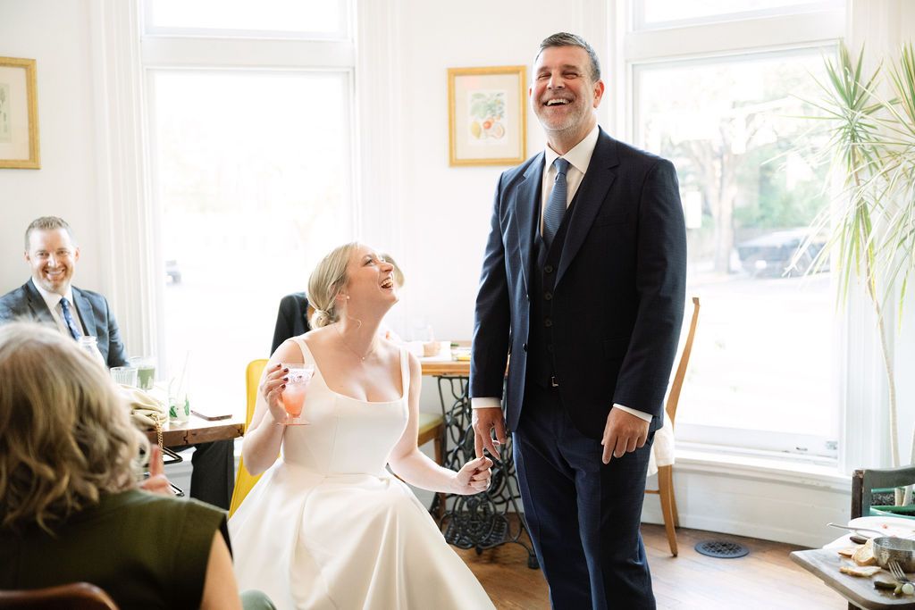 A joyful wedding moment captured in a sunlit room with bright windows and houseplants.