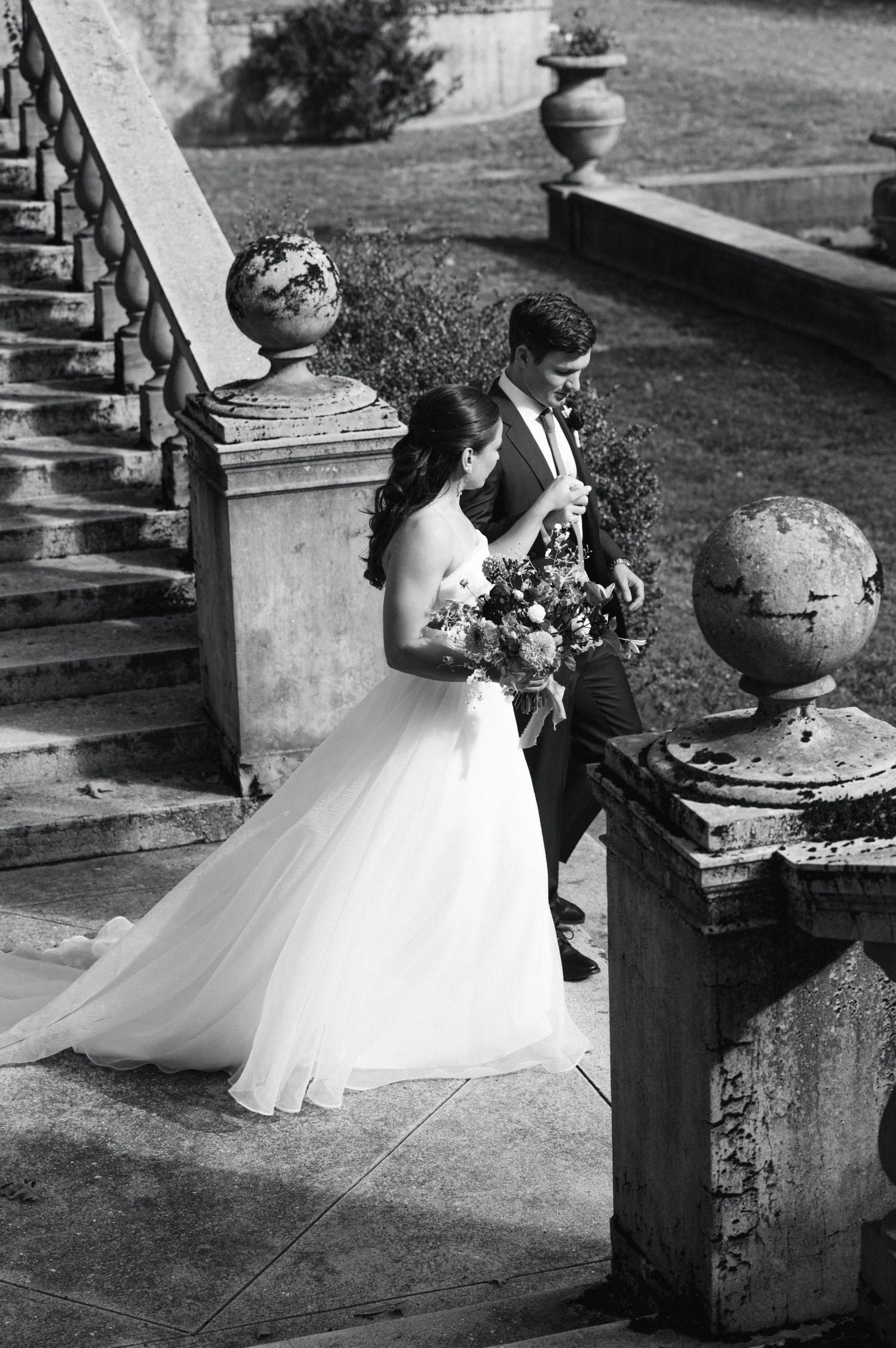 Black and white photo of bride and groom posing on ornate stone steps, bride holding bouquet in elegant wedding gown.
