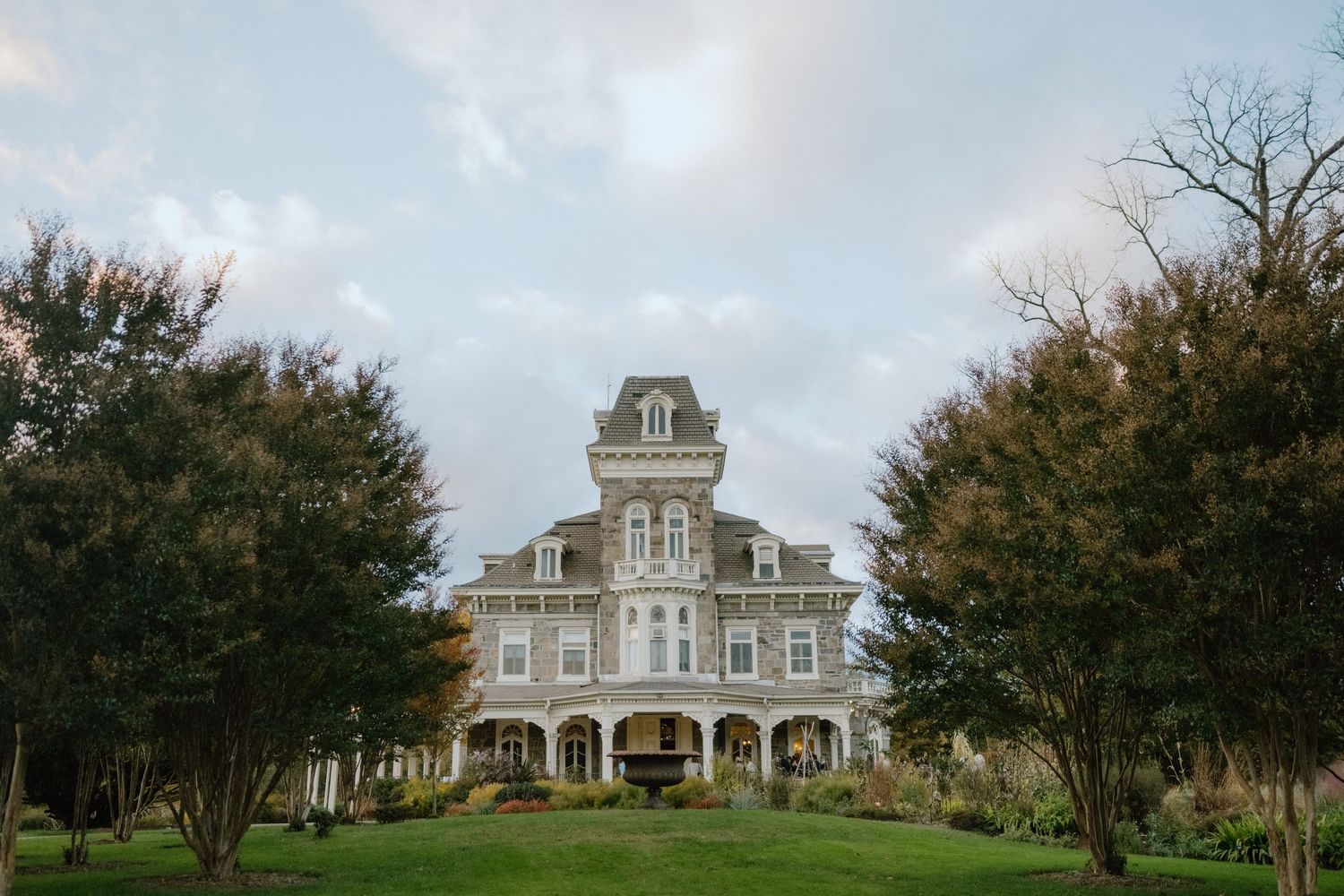 Grand Victorian mansion with wrap-around porch surrounded by lush green trees under a cloudy sky.