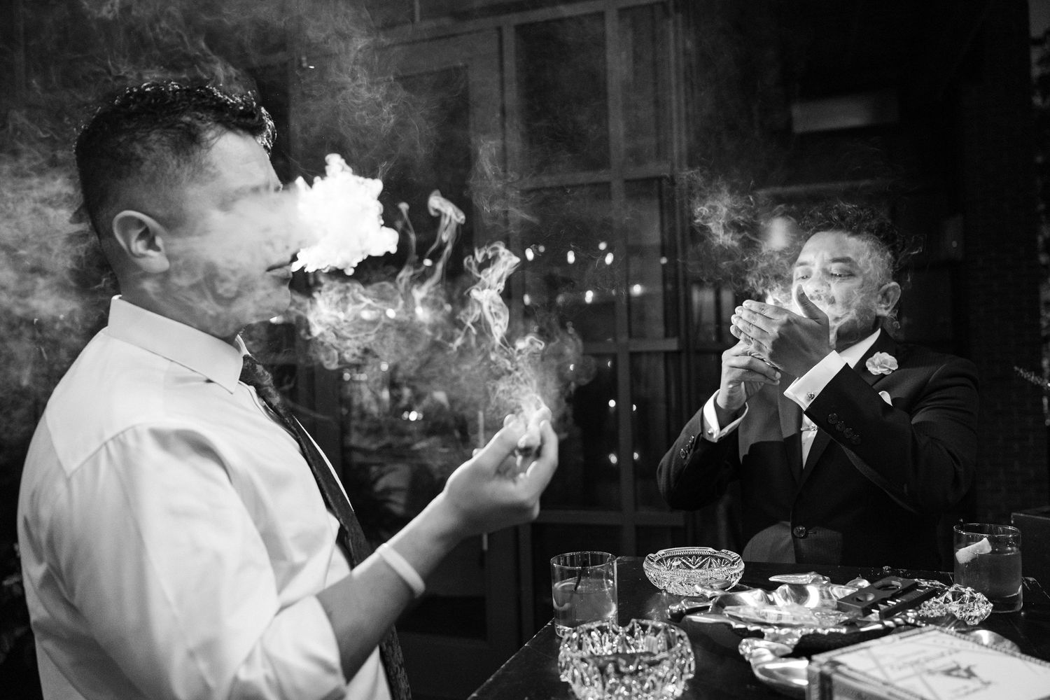 Two men in formal attire smoking cigars at a bar in a dramatic black and white wedding photo.