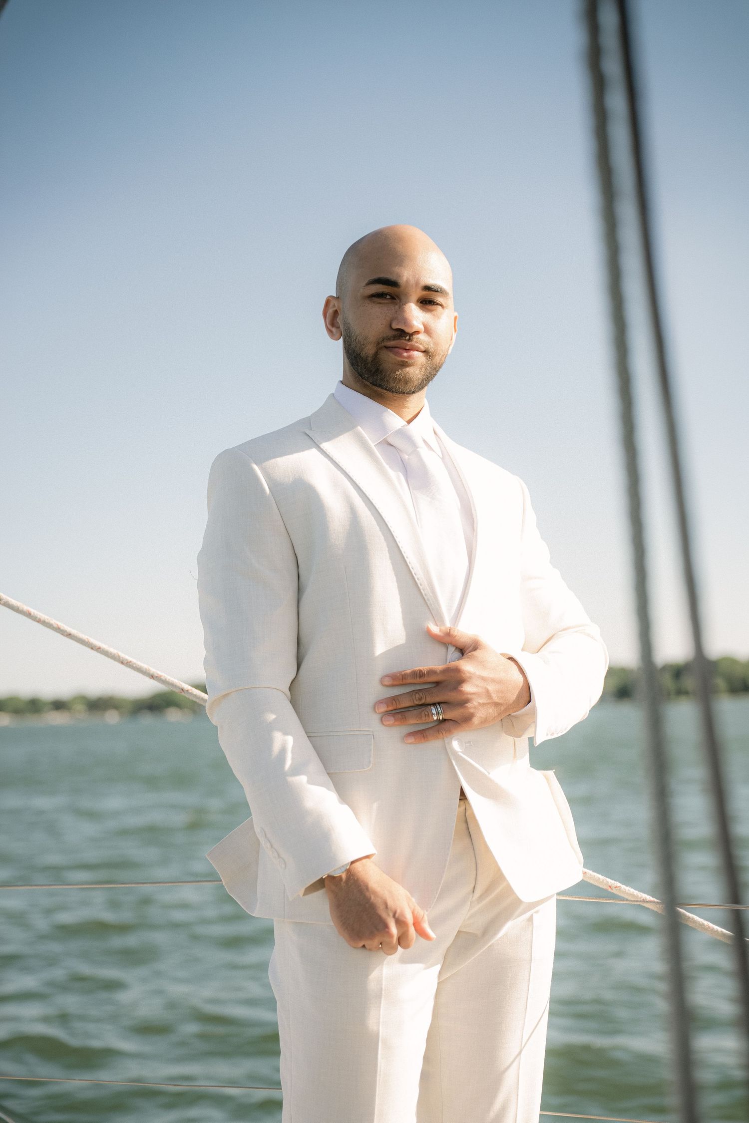 Bald man in white suit and tie standing on sailboat deck with ocean background on sunny day.