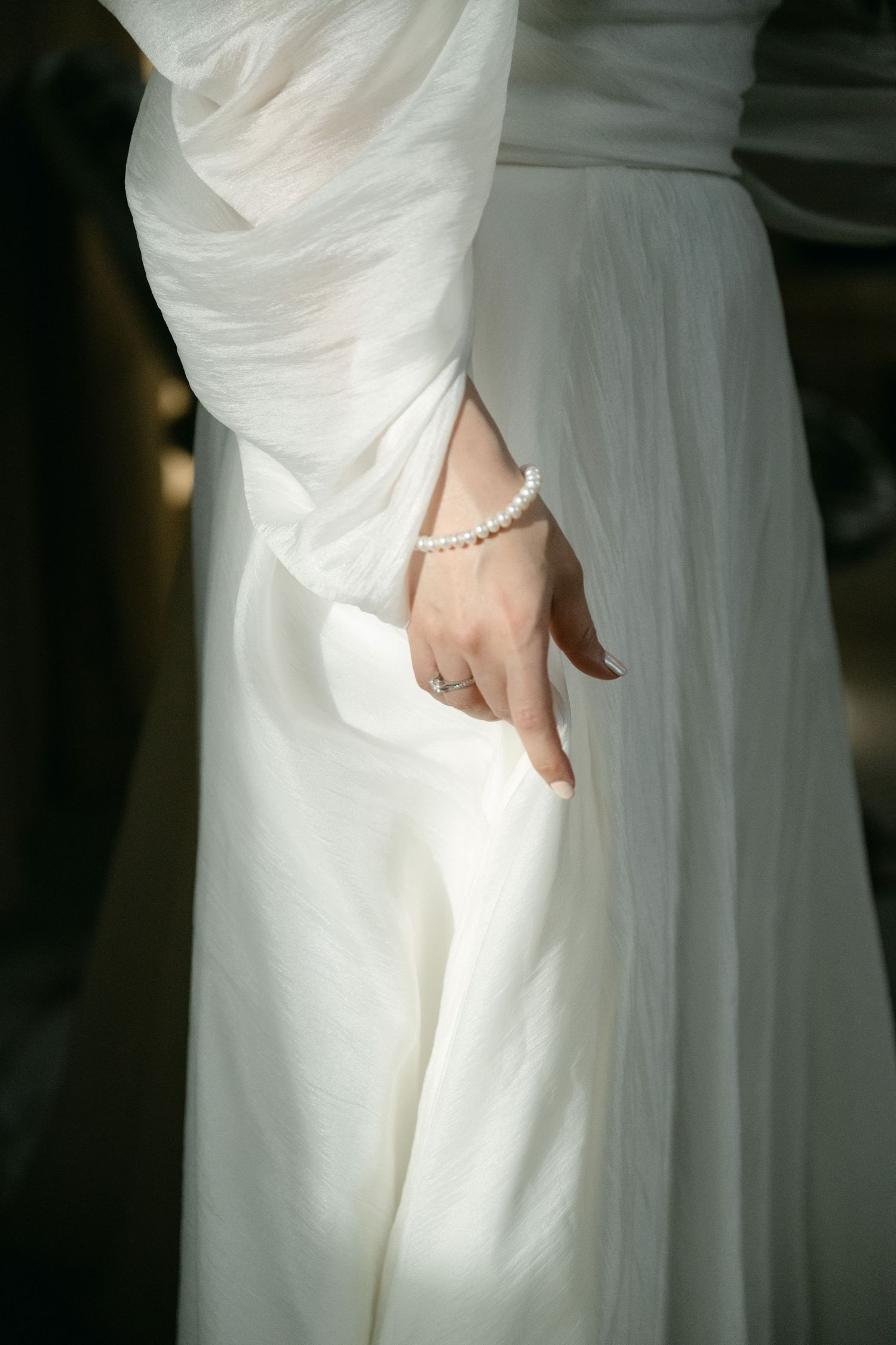 Close-up detail of a white wedding dress showing delicate fabric and a pearl bracelet.
