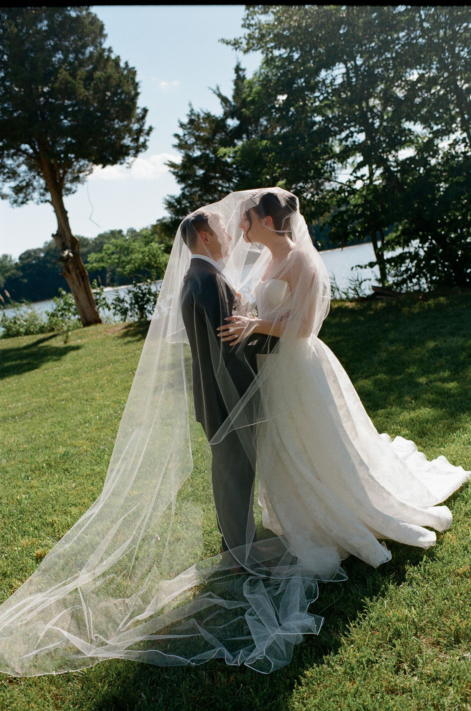 Bride and groom share a romantic moment under a flowing veil on a sunny lawn near a lake.