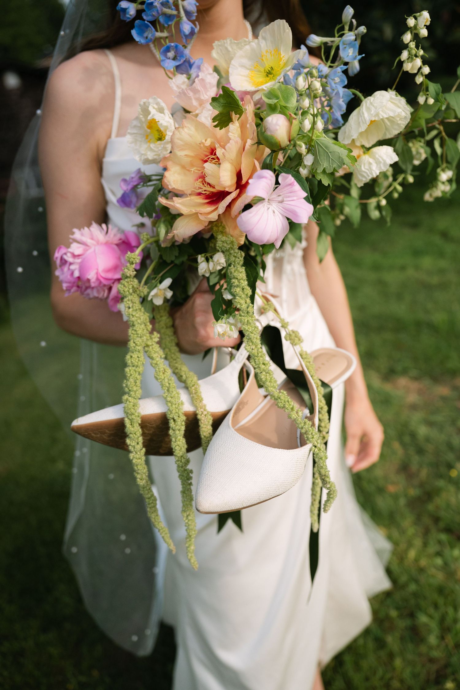 Bride in white dress holding colorful wildflower bouquet and white heels outdoors on green grass.