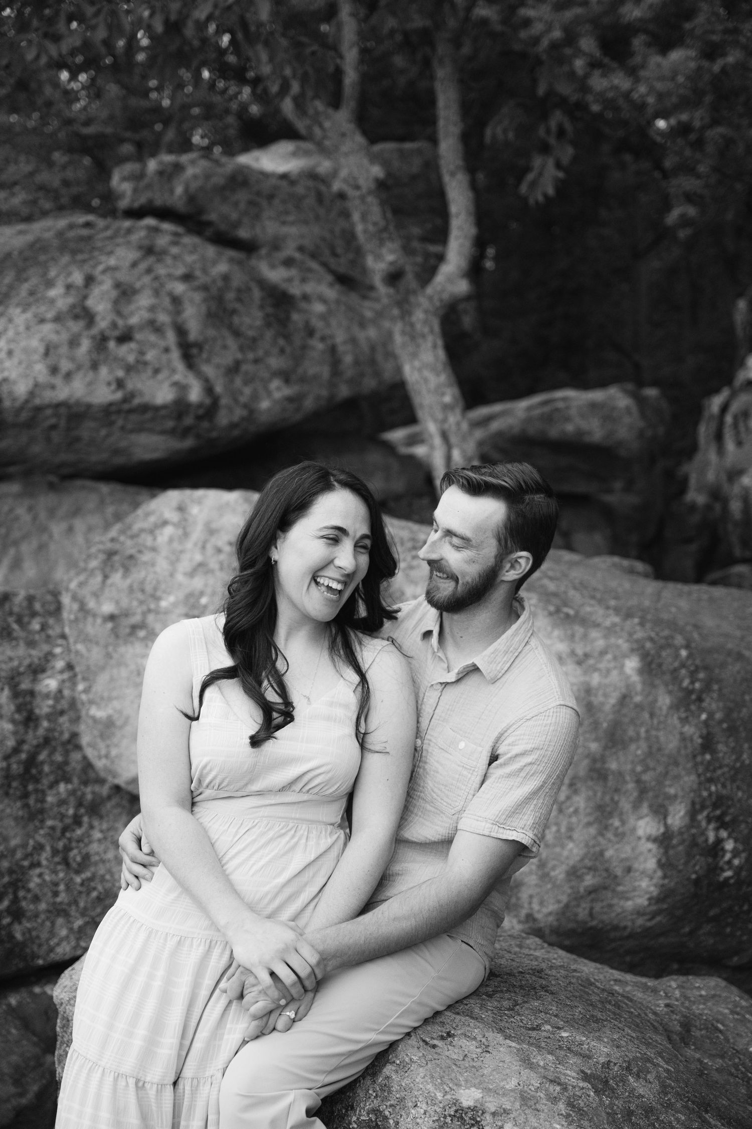 Black and white photo of a laughing couple sitting on rocks outdoors, man embracing woman in flowing dress.