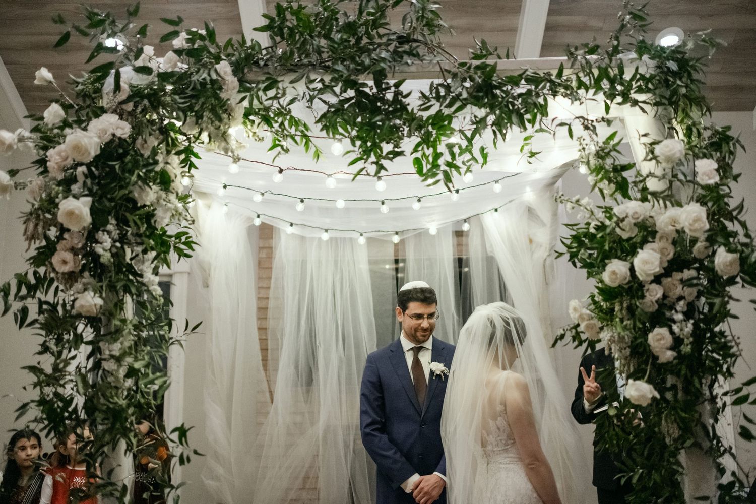 Wedding ceremony beneath an elegant floral arch with white drapery and string lights during an intimate indoor celebration.