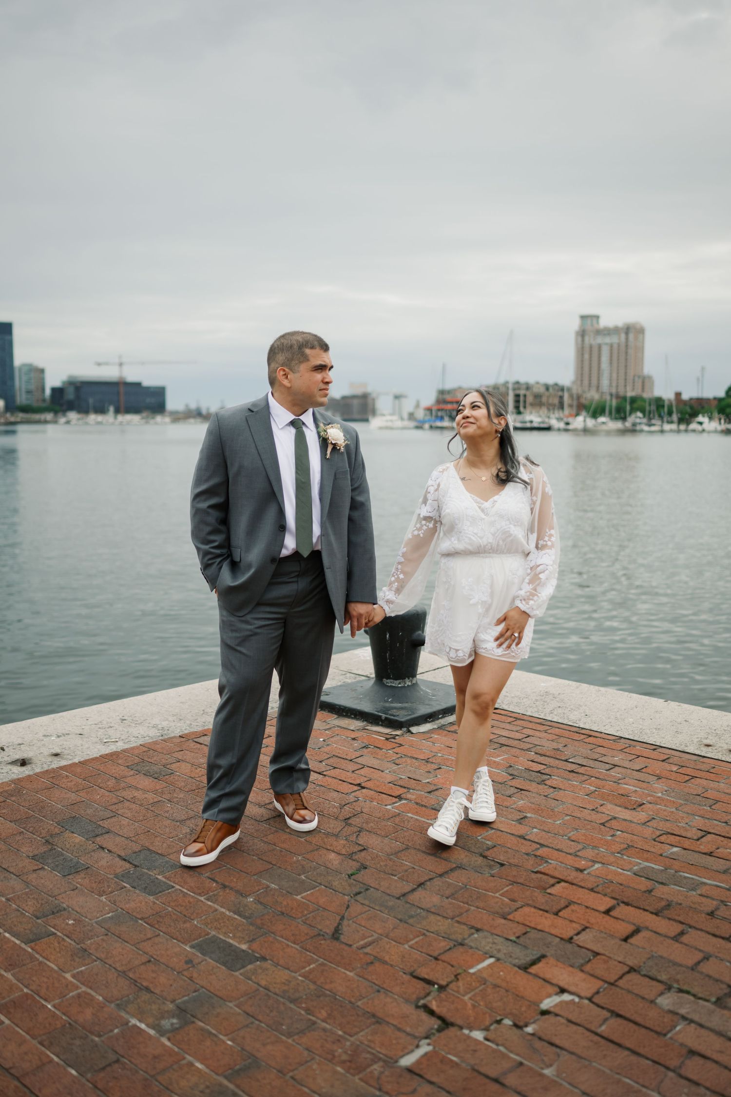 A couple shares a romantic moment on a brick pier with a harbor and city skyline in the background.
