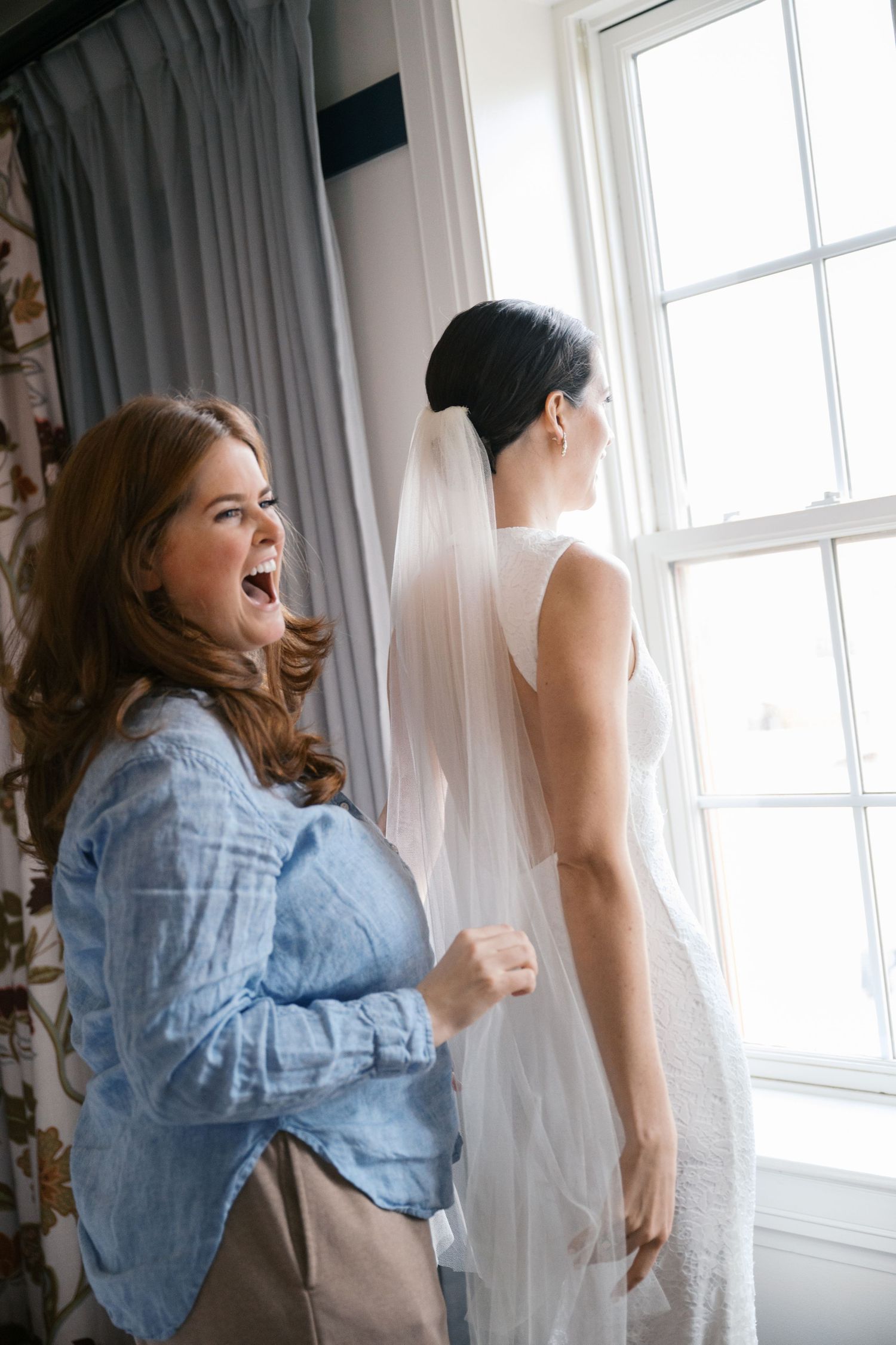 Candid moment while getting ready for a wedding, with a woman in denim reacting excitedly behind a bride in white.