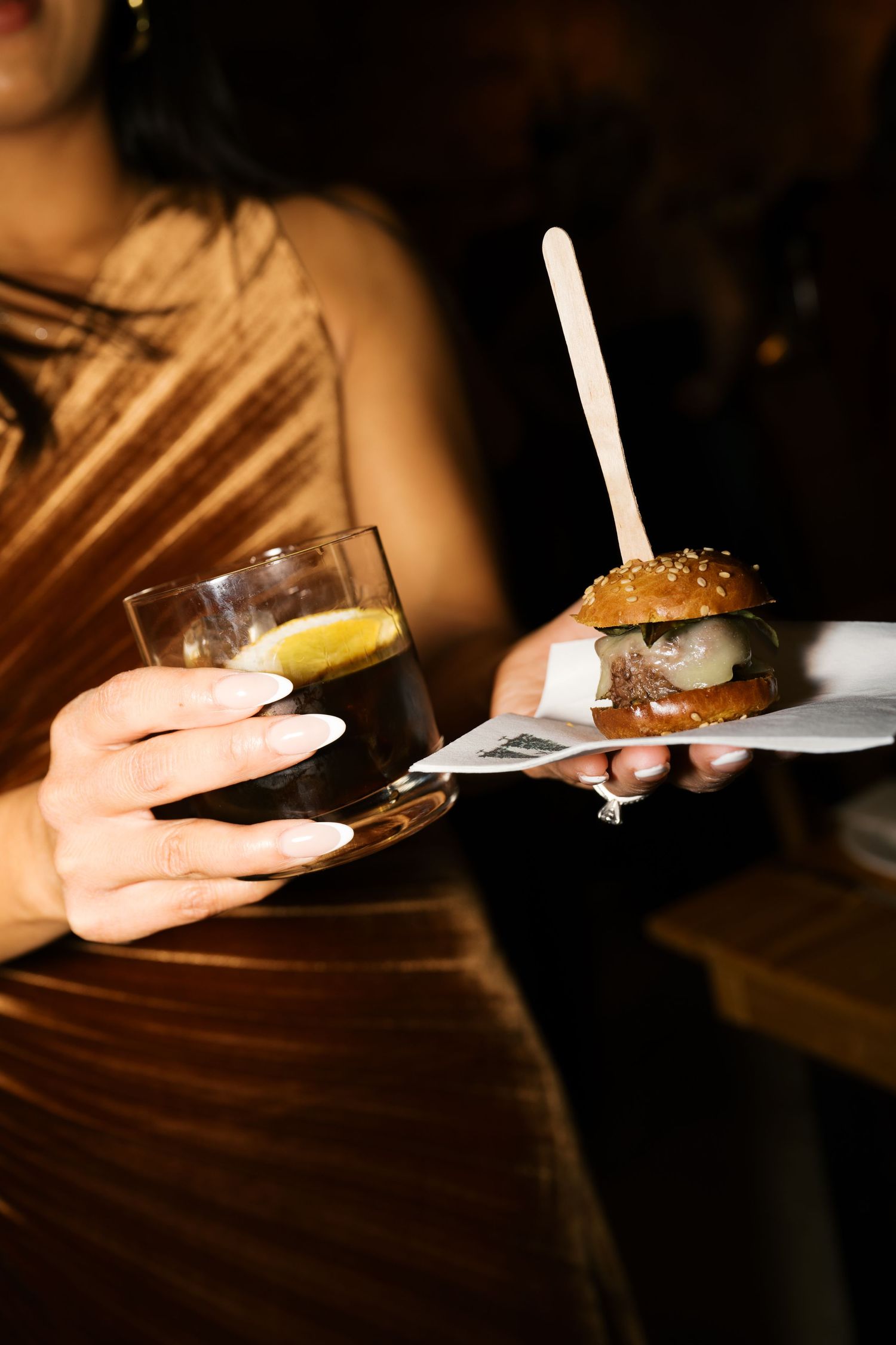 Close up of hands holding a cocktail and mini burger at an event.