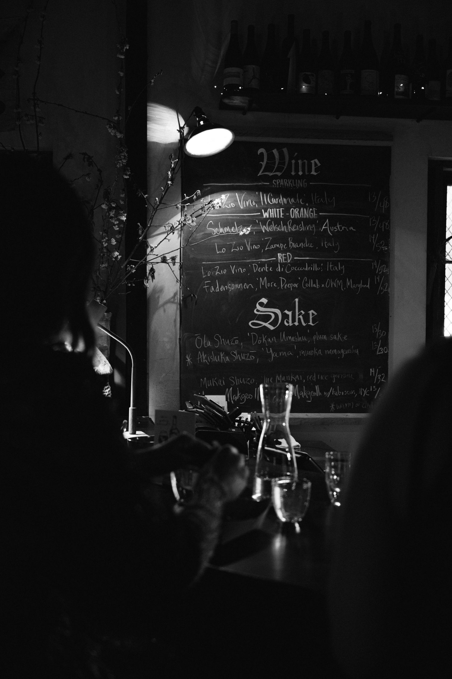A moody black and white photo of a bar menu board with ambient lighting and glassware in the foreground at Fadensonnen in Baltimore