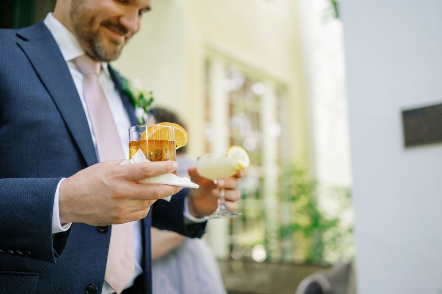Wedding guest in blue suit holds a cocktail during outdoor reception.