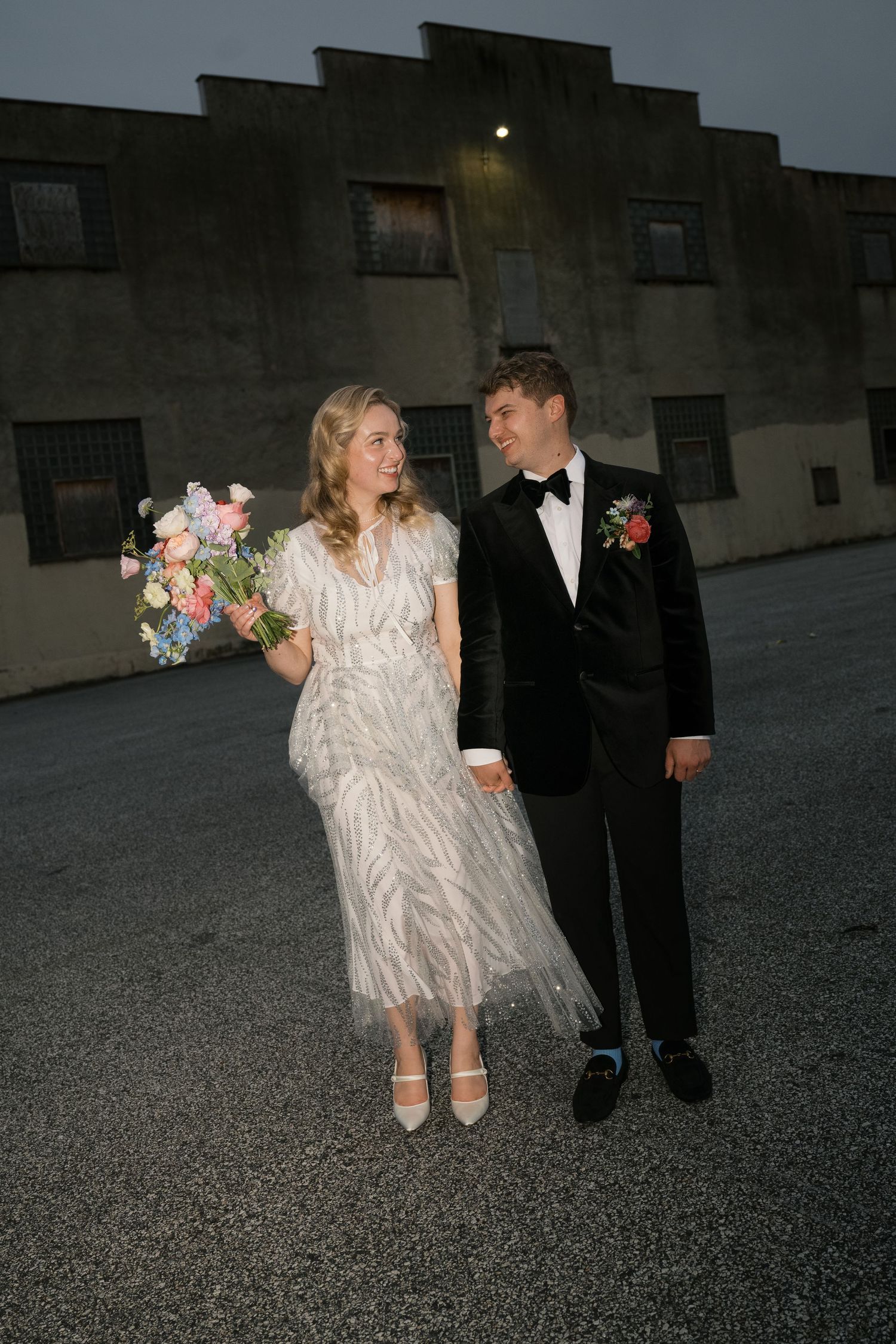 A couple in formal attire poses against an industrial building at dusk.