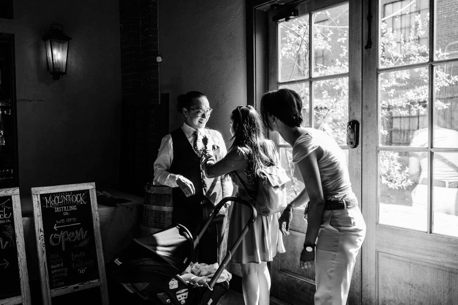 A black and white photo of a group gathering near large windows in an intimate indoor setting.