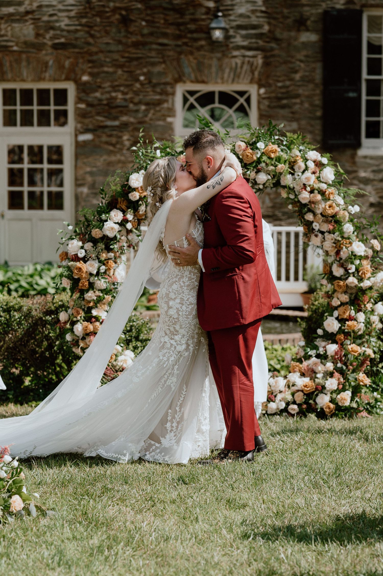 Couple shares romantic moment under floral arch with peach and white roses against stone building.