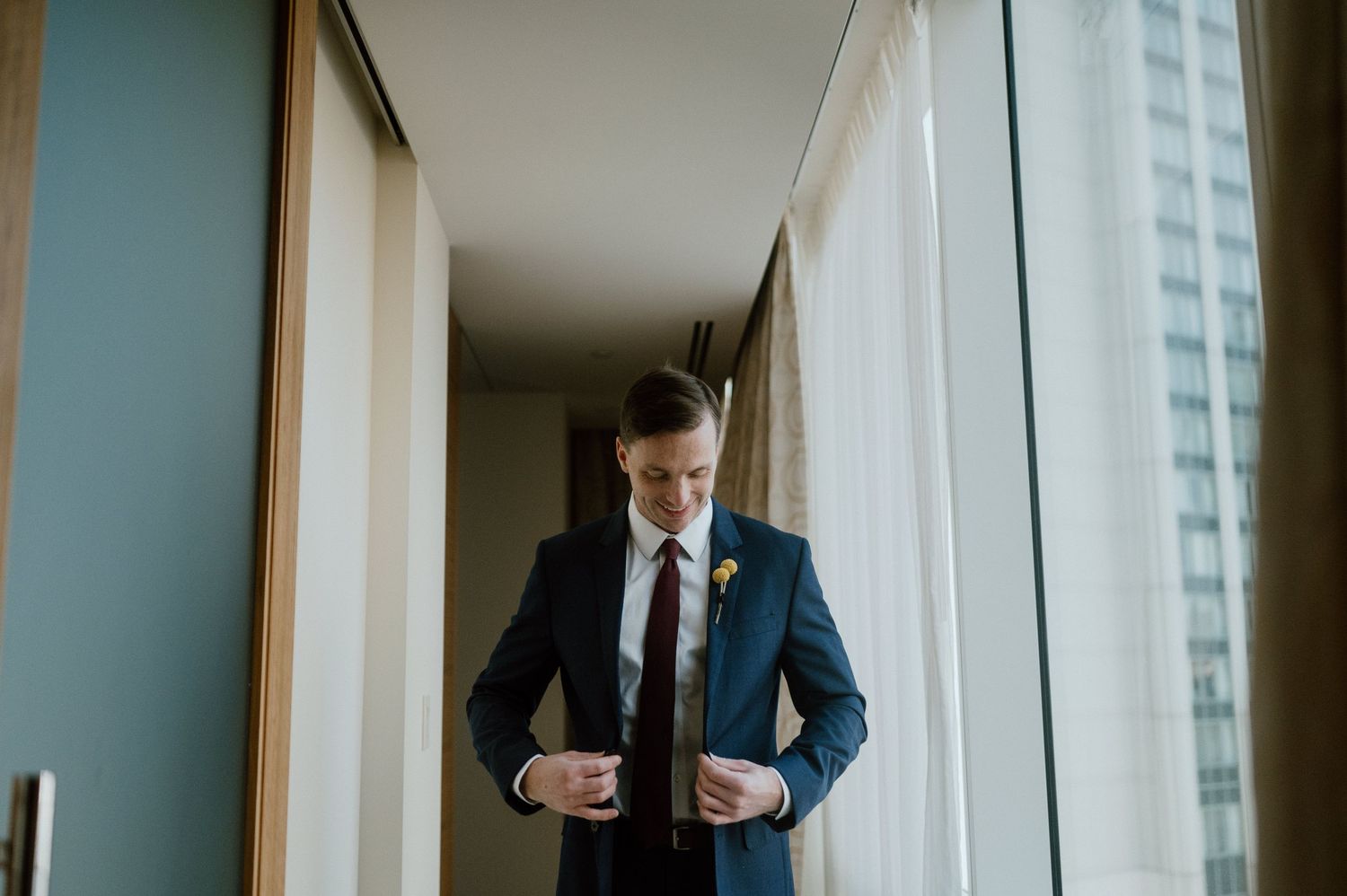 A groom in a dark suit adjusts his tie while standing near a window in a modern building.