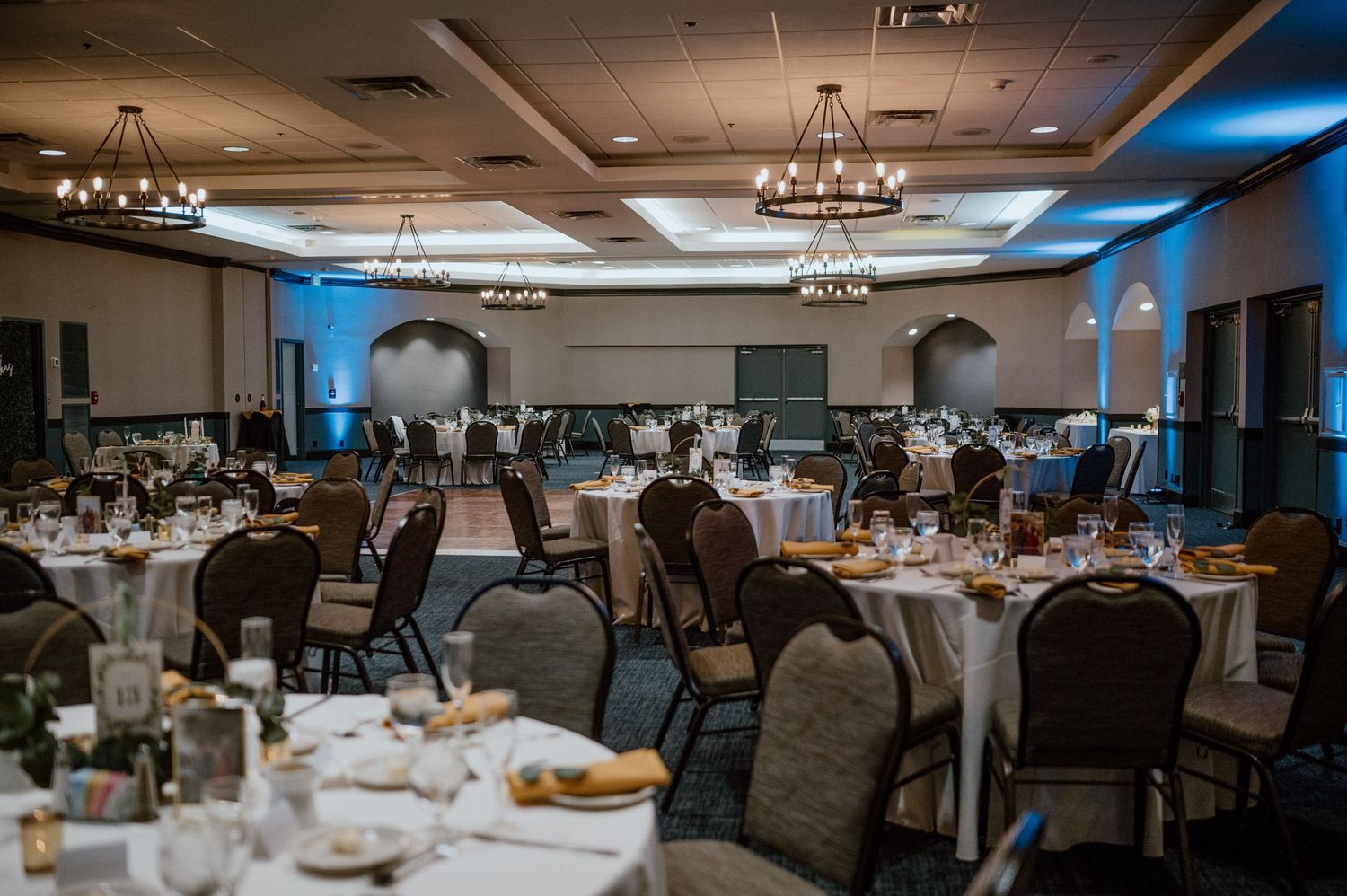 An elegant wedding reception ballroom with round tables, chairs and chandeliers set up for dining.