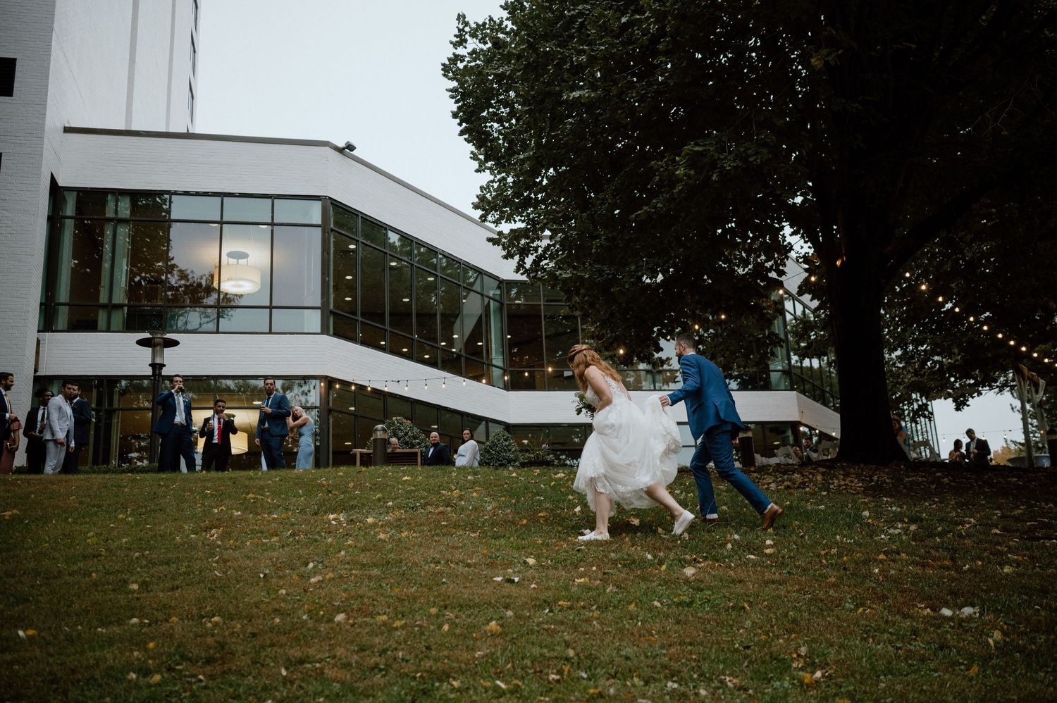 Wedding couple running playfully across lawn with white dress flowing in wind near modern building.