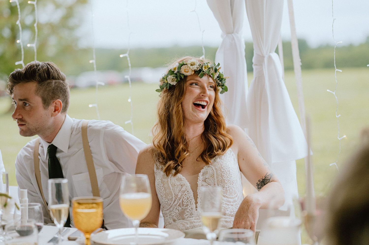 Joyful candid moment during wedding reception under white tent with floral crown and champagne glasses.