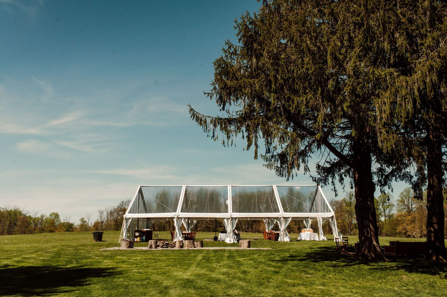 Clear outdoor wedding tent setup on a lush green lawn with tall evergreen trees in the background.