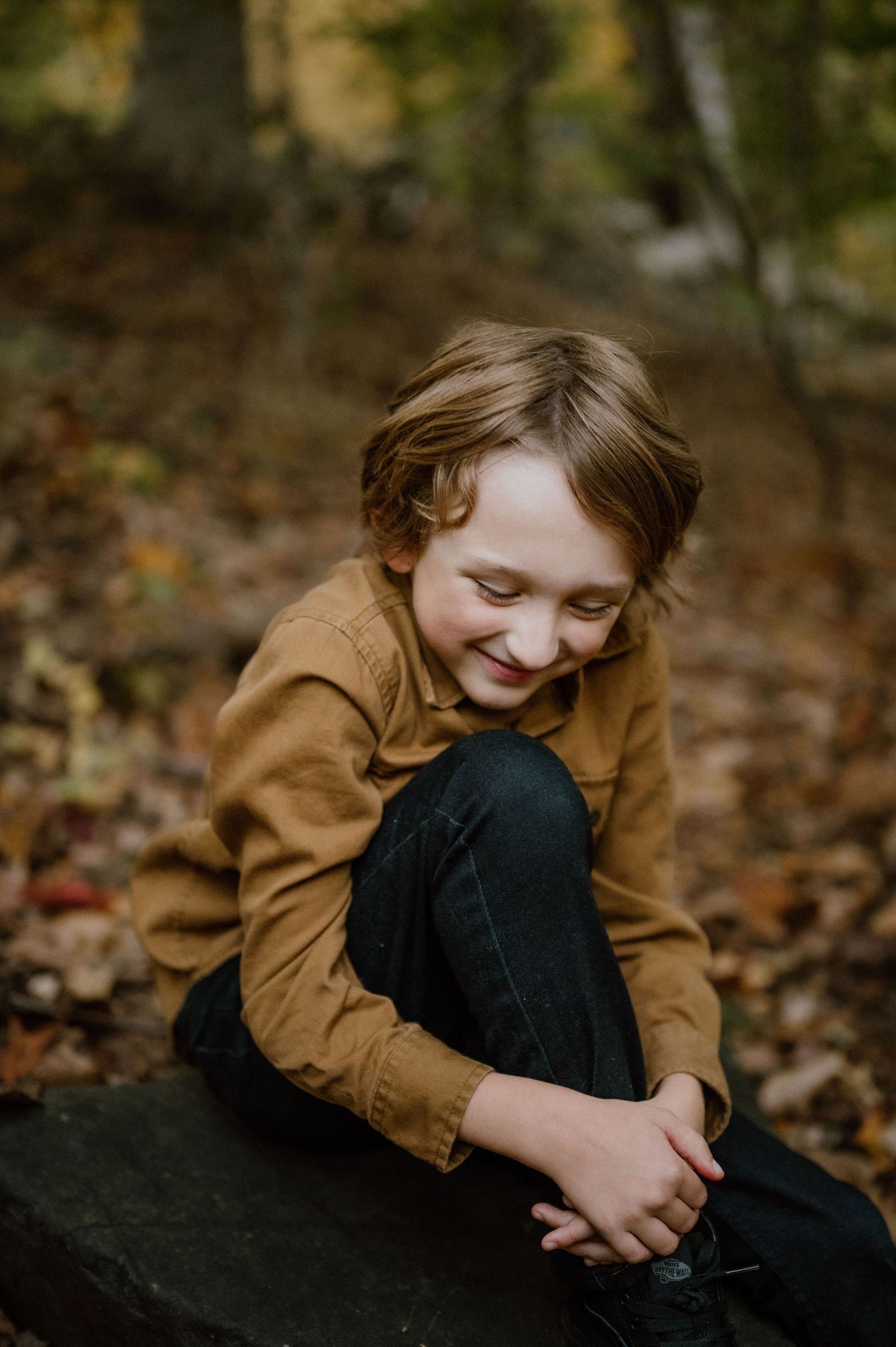 Child in brown jacket sits contemplatively on a rock surrounded by fallen leaves in the woods.