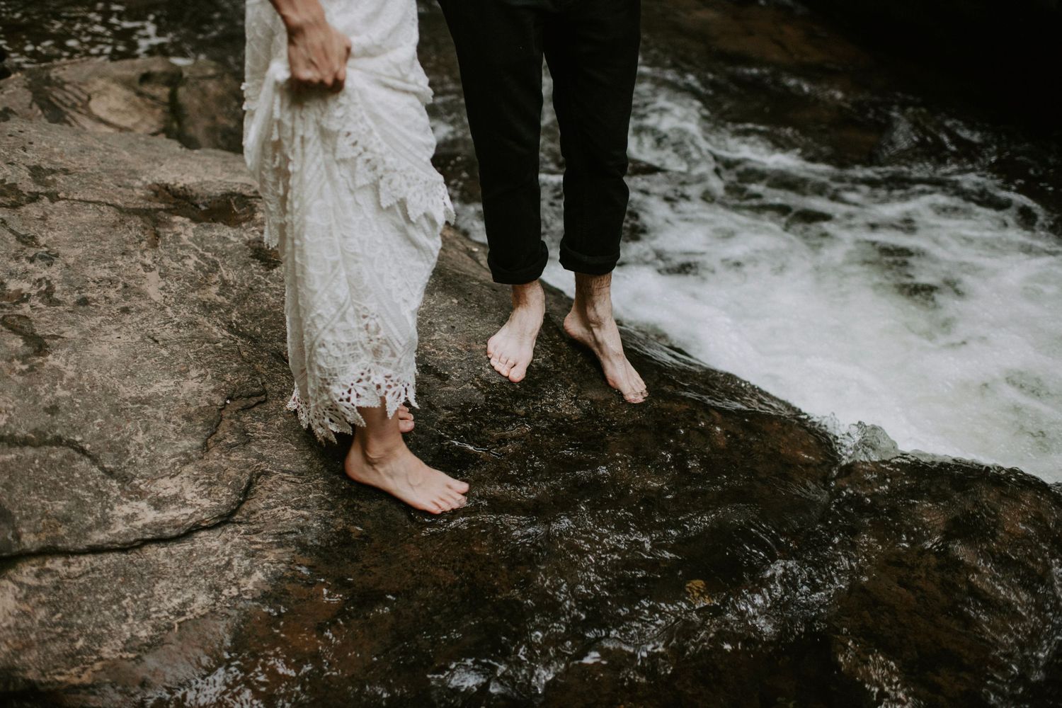 Barefoot couple standing on wet rocks by rushing stream, showing just their feet and lower legs.