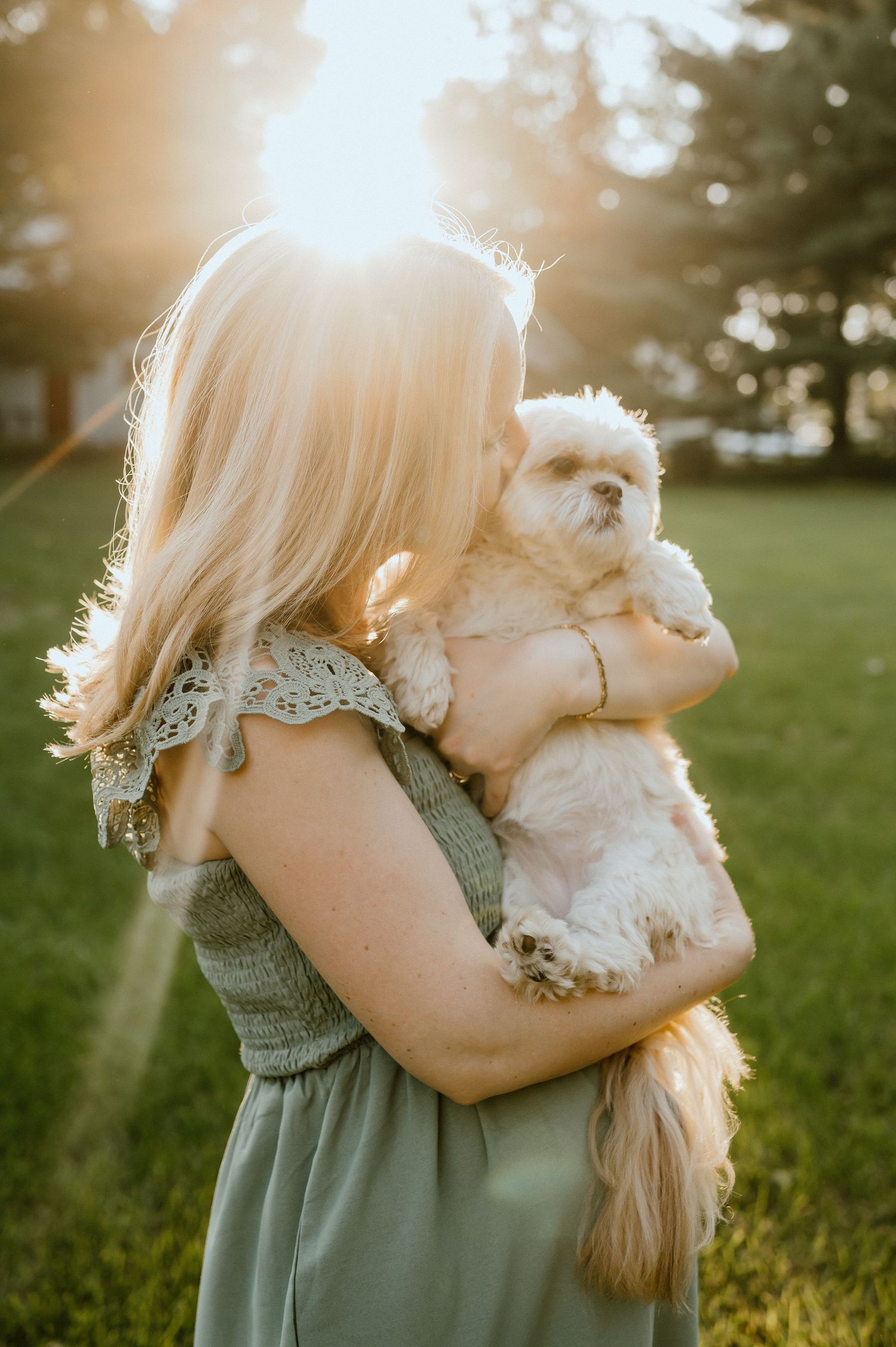 Woman in sage dress holds small white dog against golden sunset backdrop.