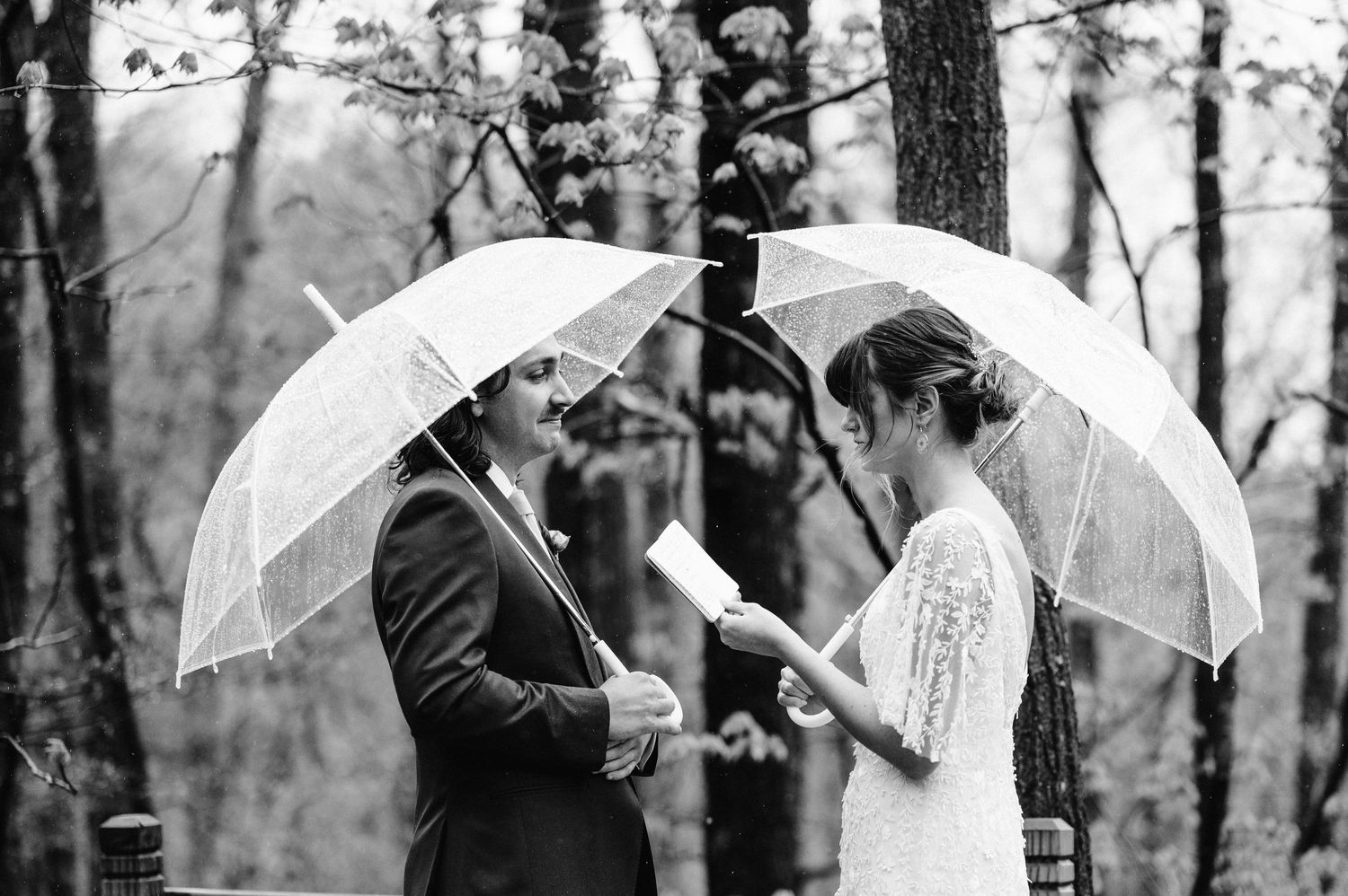 A couple shares a moment under white umbrellas during their rainy forest wedding ceremony.