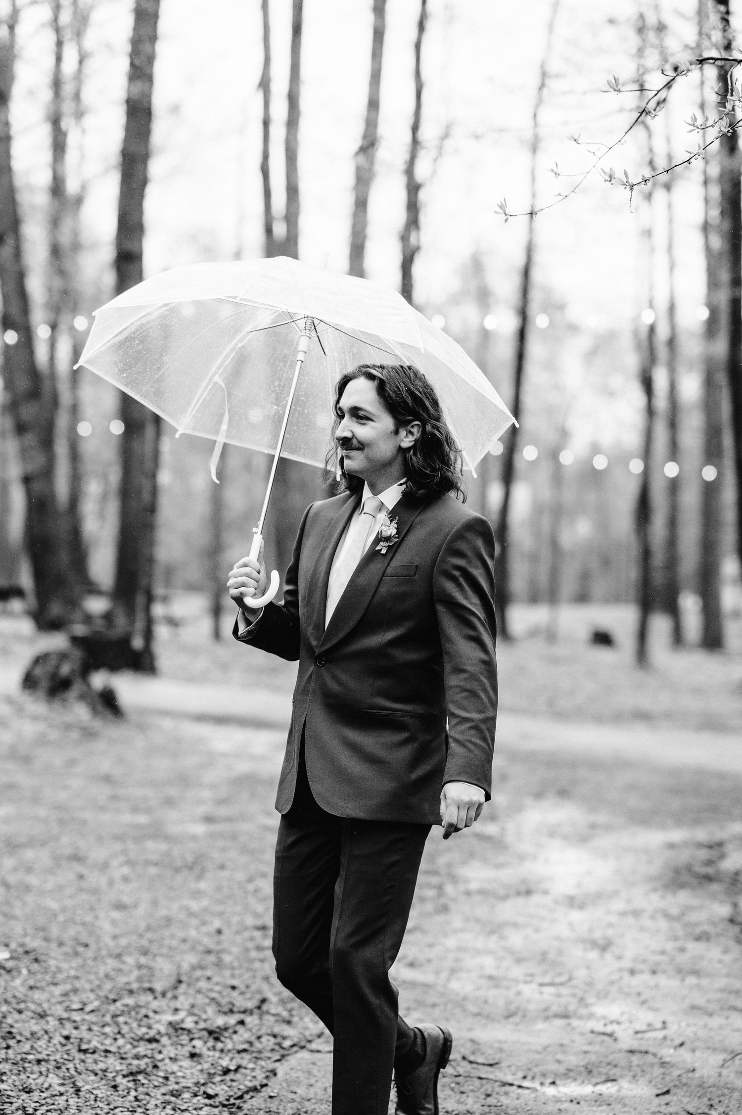Figure in suit holding clear umbrella stands on forest path with string lights in background.