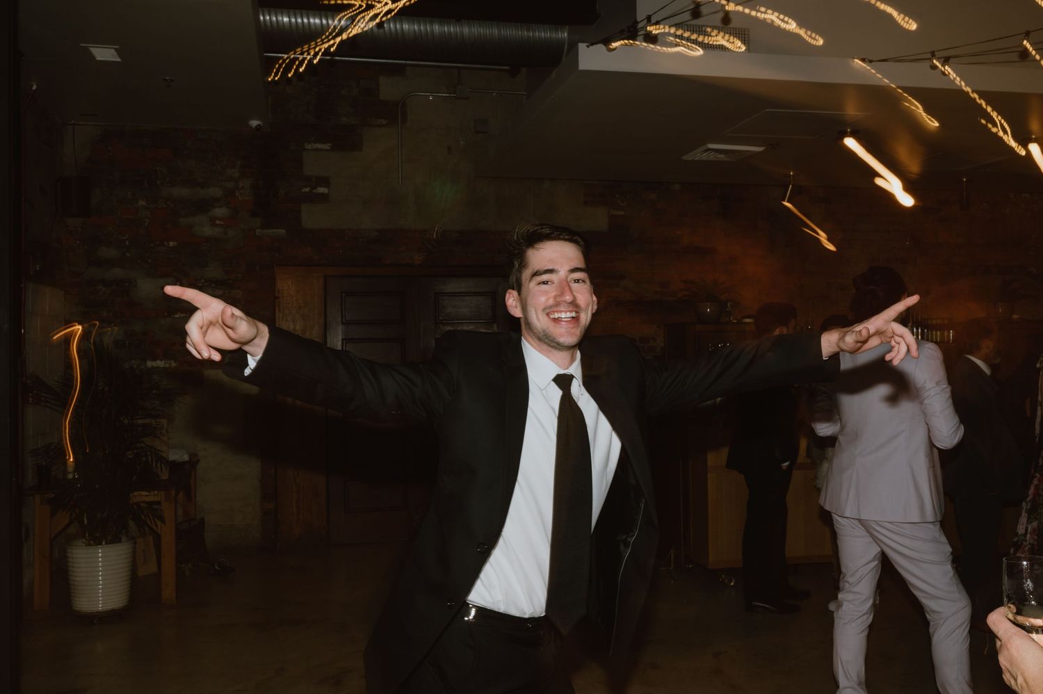 A guest celebrates on the dance floor with arms spread wide at a dimly lit wedding reception.