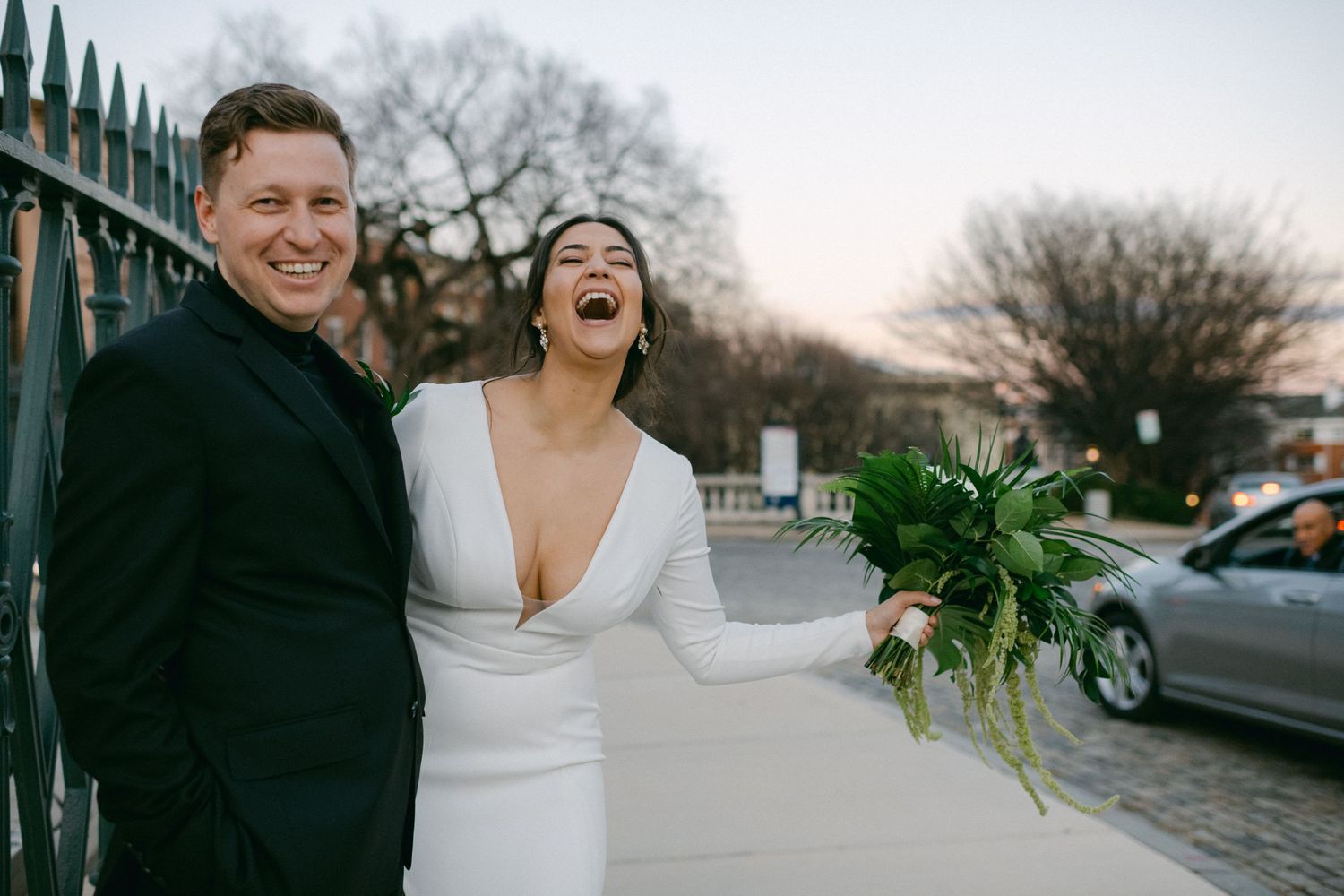 Joyful wedding couple laughing together outdoors with white bouquet.