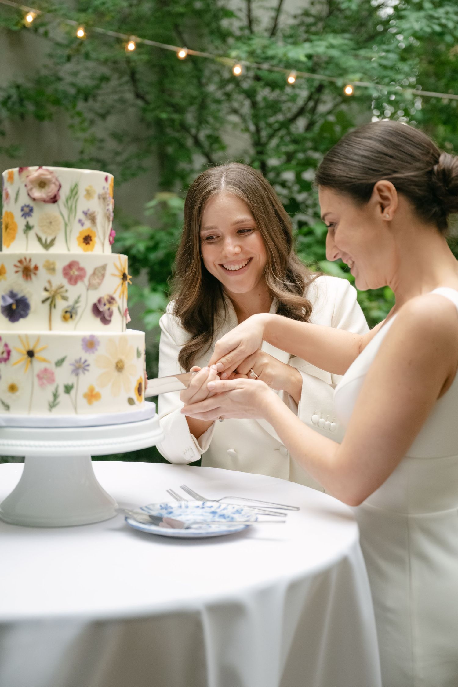 Three floral pressed wedding cakes displayed on white tablecloths with soft string lights and greenery in background.
