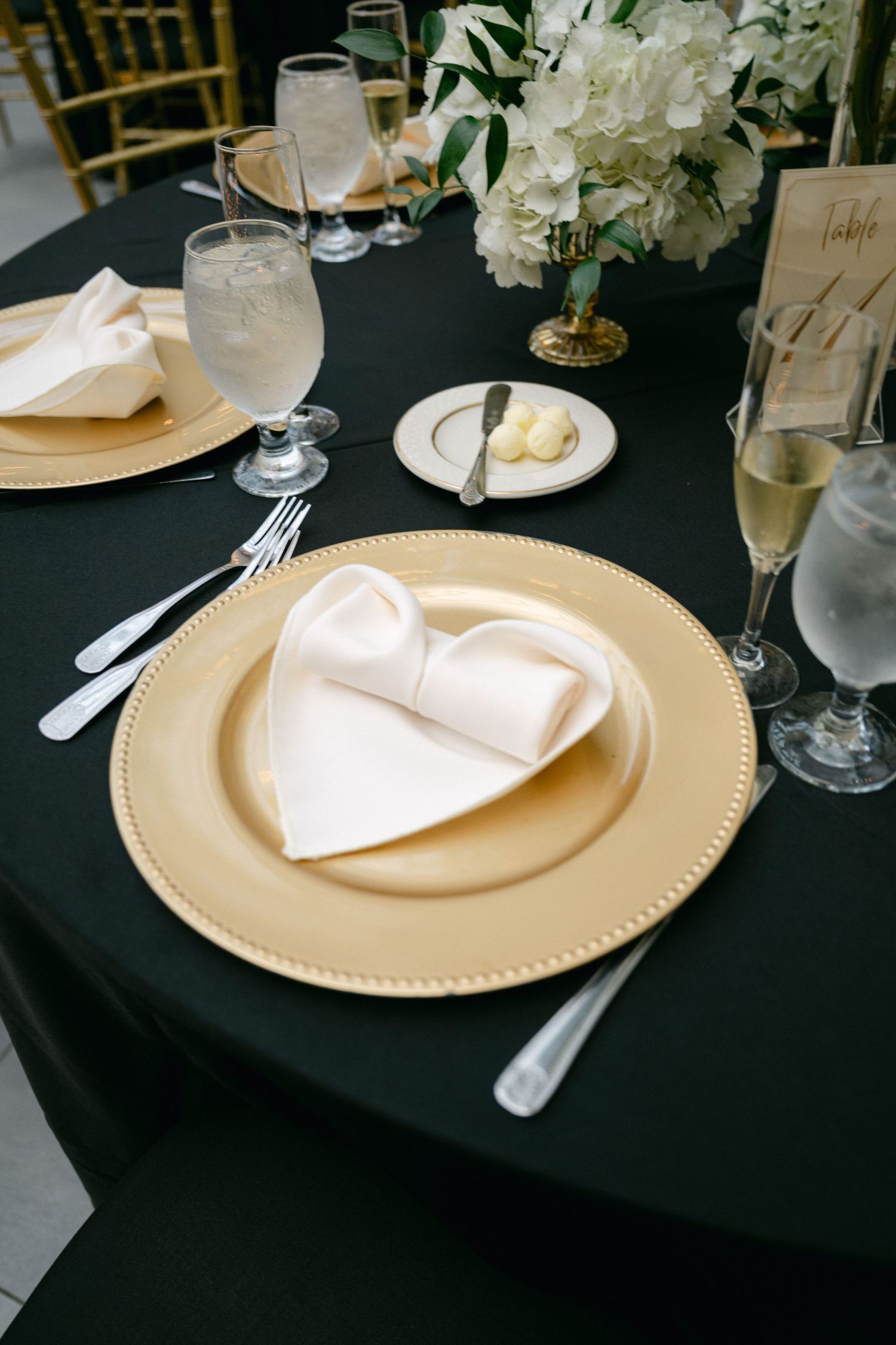 Elegant place setting with gold charger plate, white napkin, and floral centerpiece on black tablecloth.