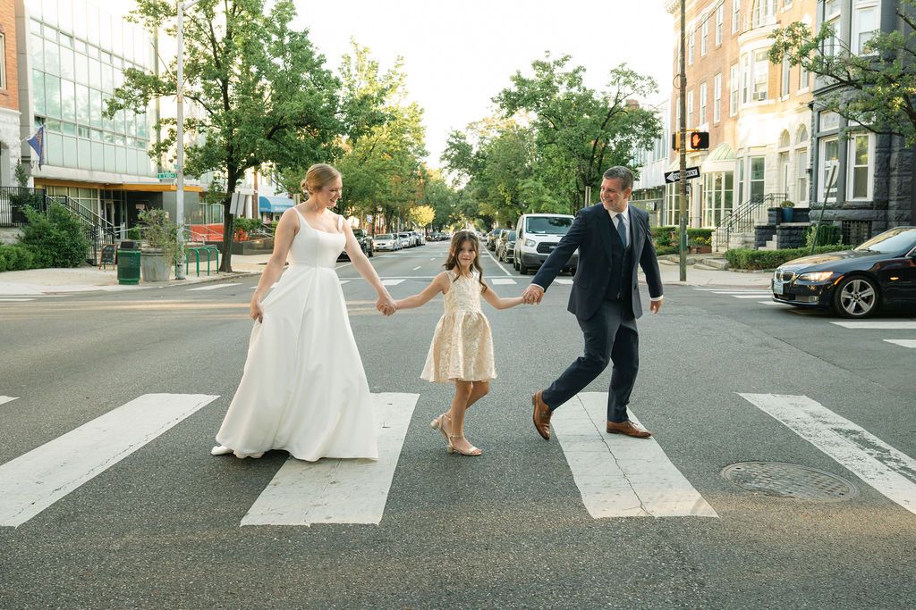 Newlyweds walking across a crosswalk on a tree-lined city street.