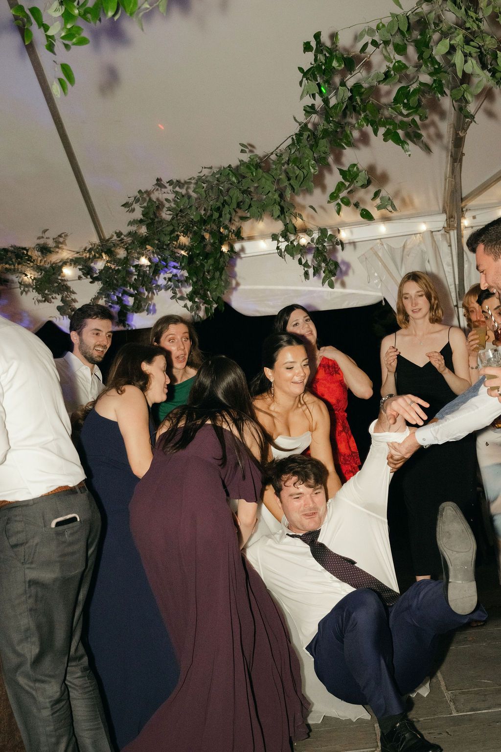 Wedding reception guests dance enthusiastically under a tent decorated with hanging greenery.