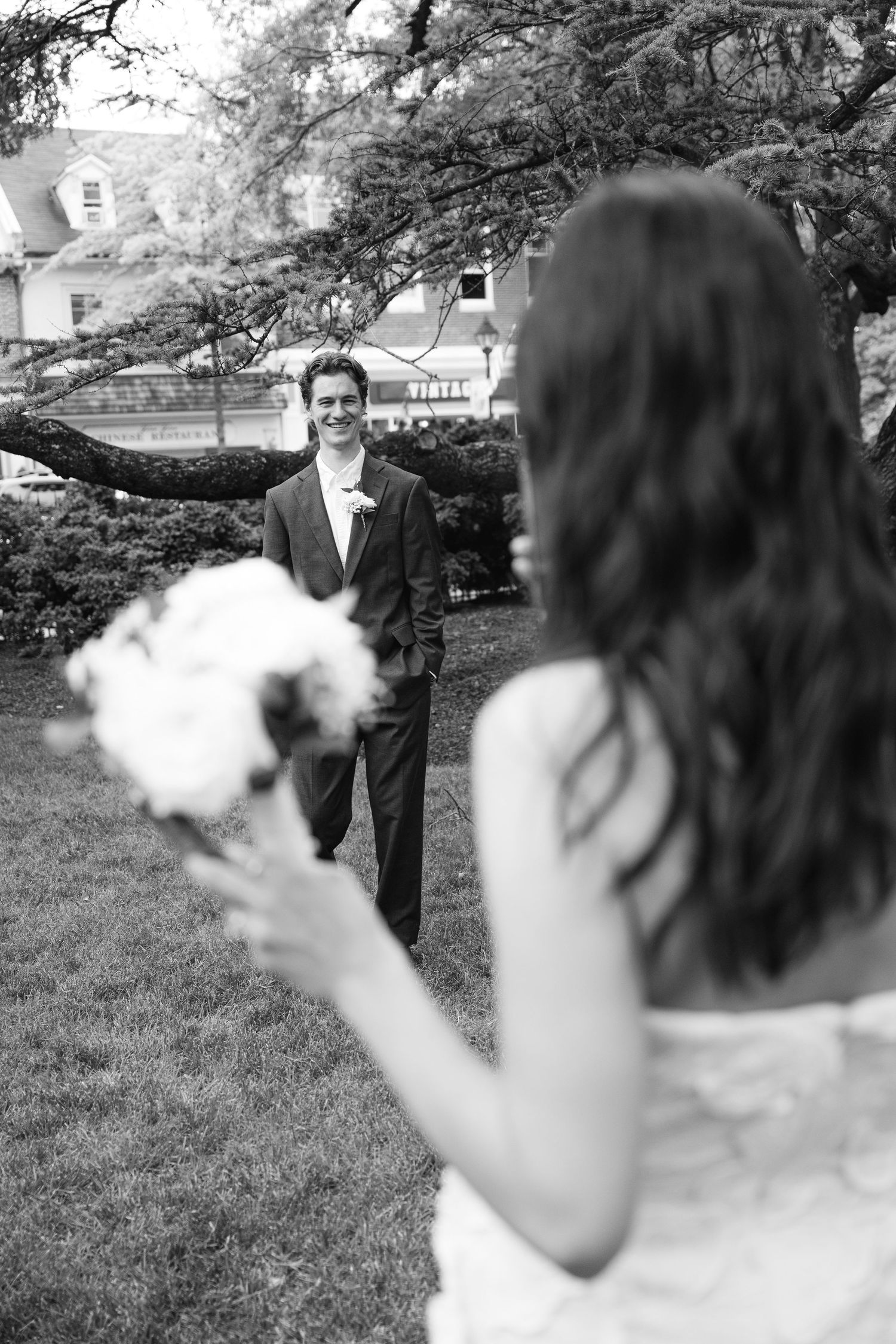 Black and white photo of a smiling groom seeing his bride in a wedding dress holding a bouquet outdoors.
