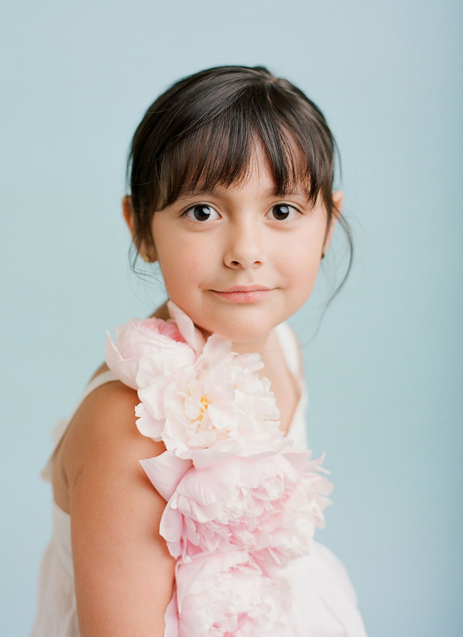 little girl with black hair dark brown eyes wearing real peonies attached to her sundress against soft blue backdrop