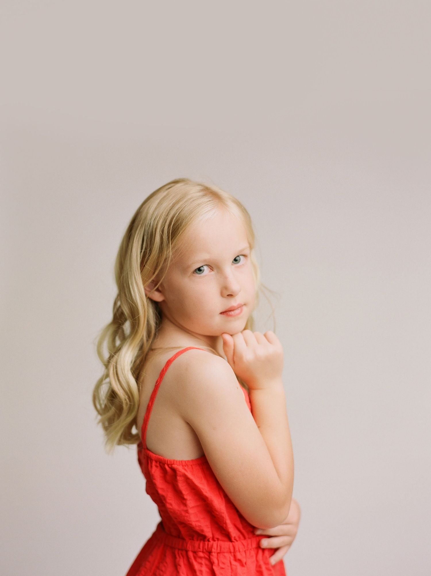 little girl with blonde hair/blue eyes looking over her shoulder at camera against neutral backdrop
